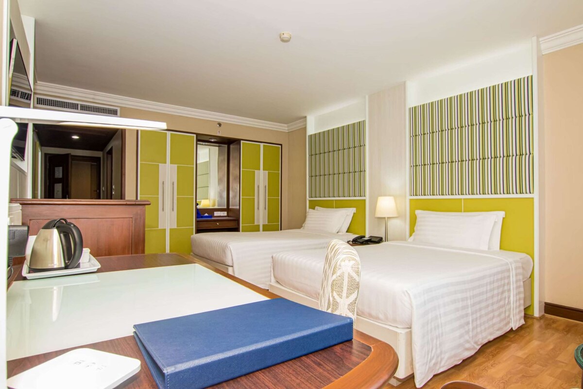 Habitación de hotel con dos camas, un escritorio con hervidor, un espejo y una ventana con persianas verticales.