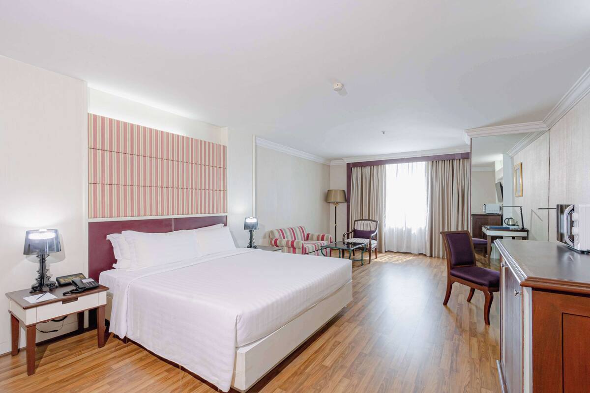 A hotel room with a large bed, a desk with a lamp, a chair, a sofa, and a dining area with a table and chairs.