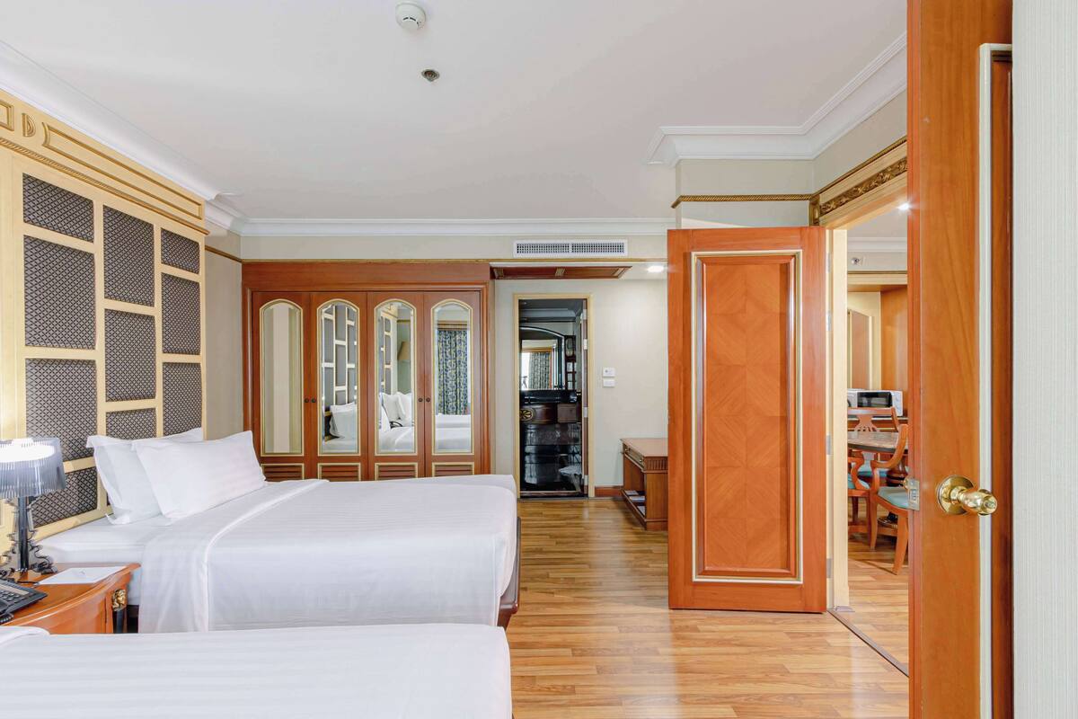 A hotel room with two beds, wooden flooring, a desk with a lamp, and a dining area with chairs and a table.