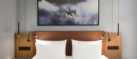 A modern hotel room with a large bed, a headboard with integrated lighting, bedside tables, and a framed artwork above the bed.