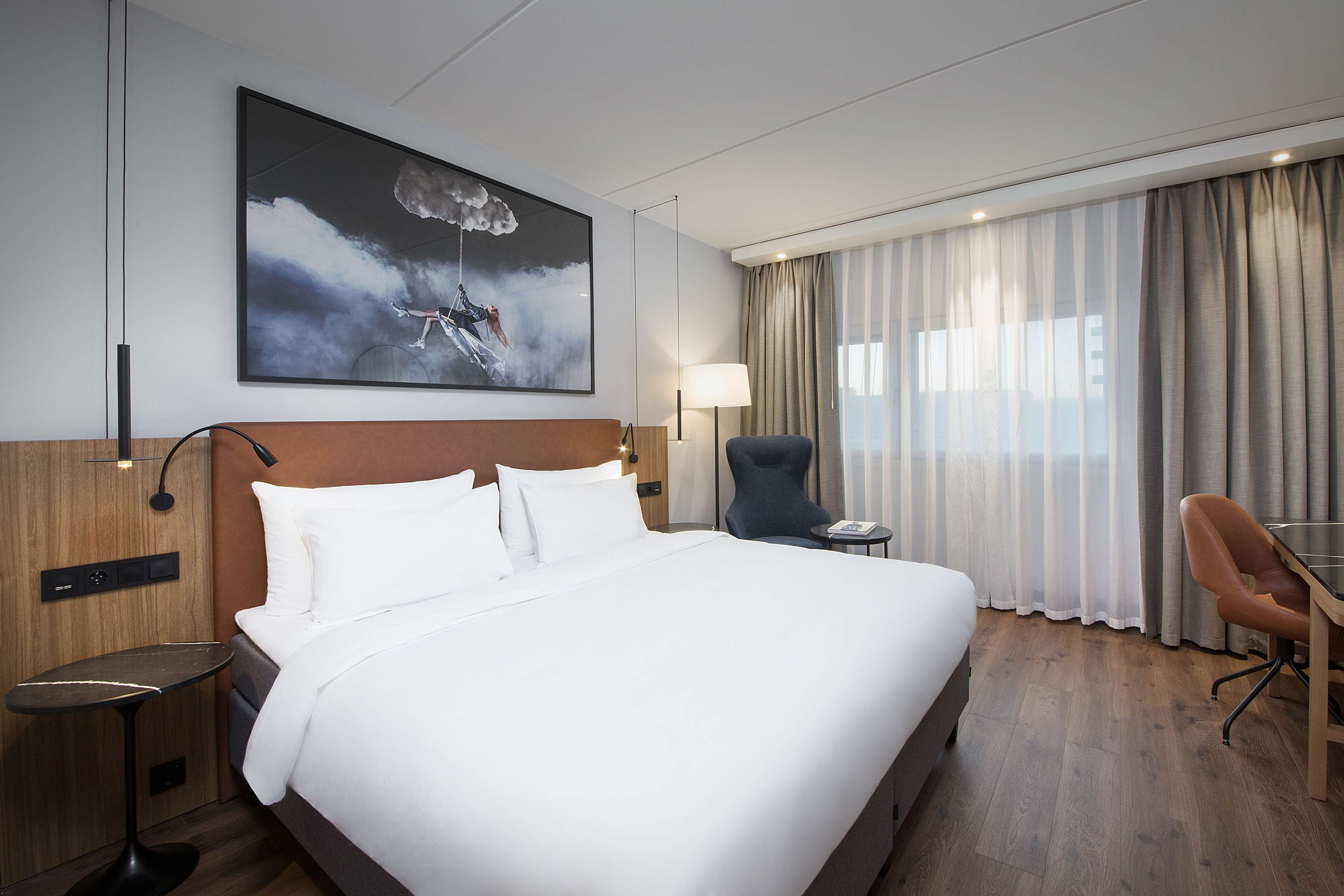 A hotel room with a large bed, a desk, a chair, a lamp, and a painting on the wall.