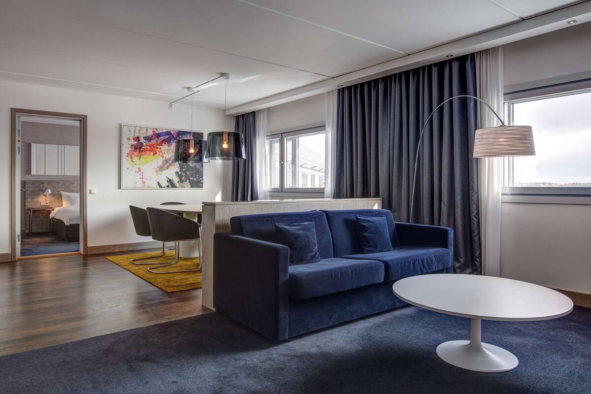A modern hotel room with a dark blue sofa, a white round table, and a large window with curtains.