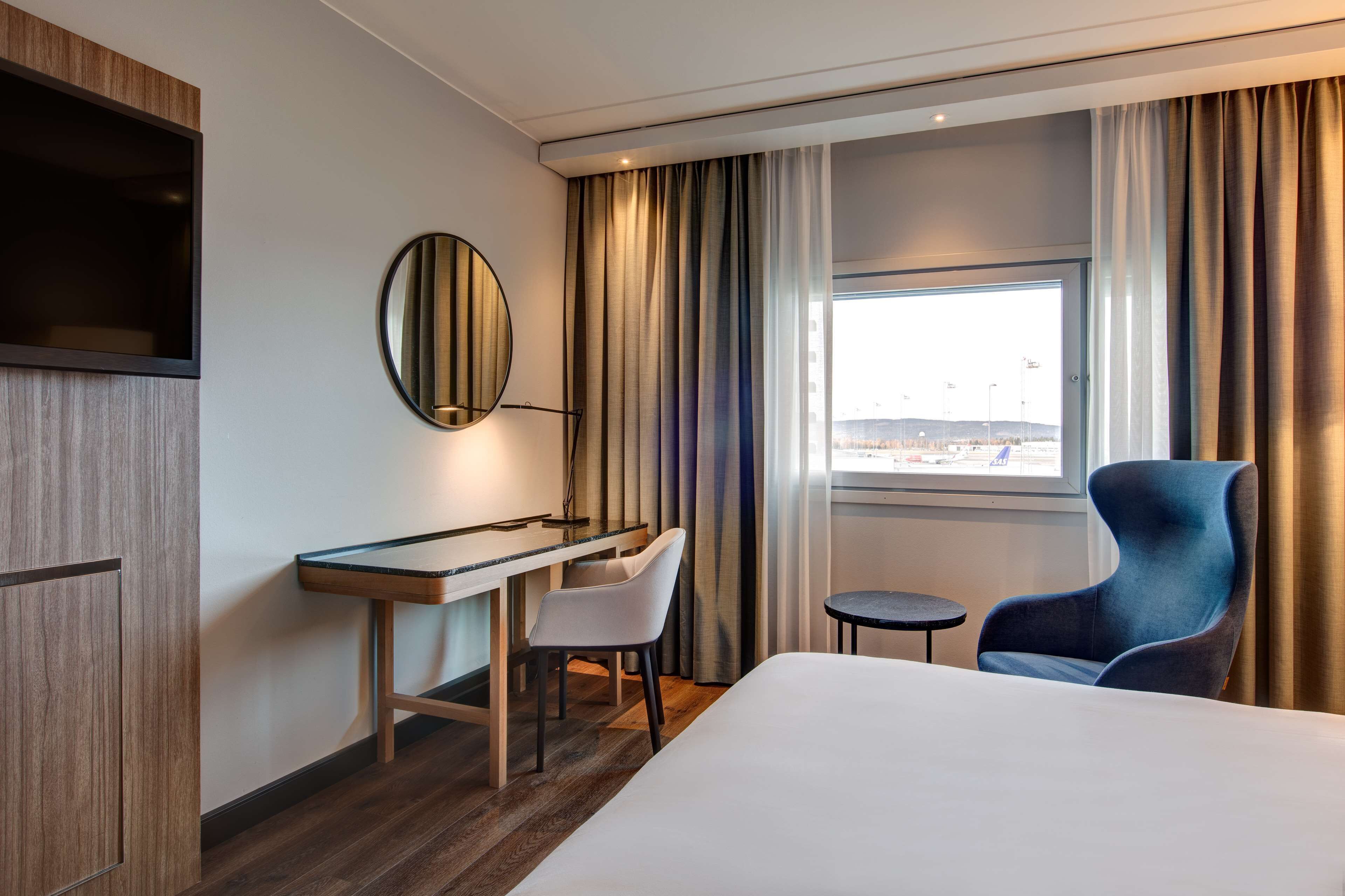 A hotel room with a bed, a desk, a chair, a small table, a mirror, and a window with curtains.