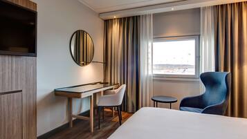 A hotel room with a bed, a desk, a chair, a small table, a mirror, and a window with curtains.