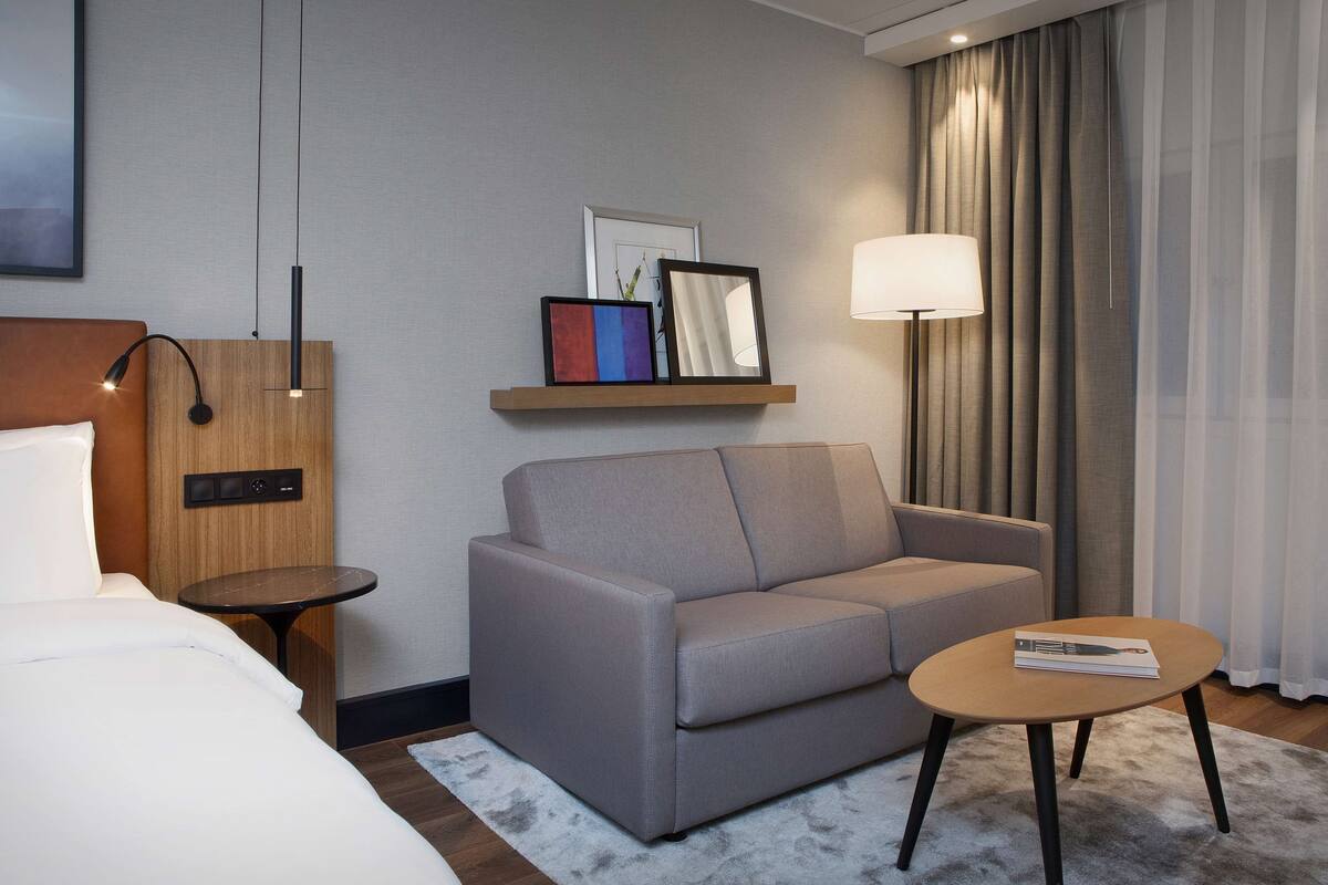A modern hotel room with a grey sofa, a small round table, a bedside table, a lamp, and a shelf with framed pictures.
