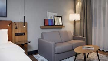 Une chambre d’hôtel moderne avec un canapé gris, une petite table ronde, une table de chevet, une lampe et une étagère ornée de tableaux encadrés.