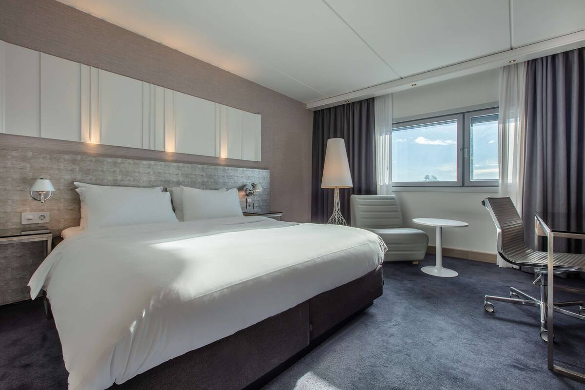 A modern hotel room with a large bed, a desk, a chair, and a window with curtains.