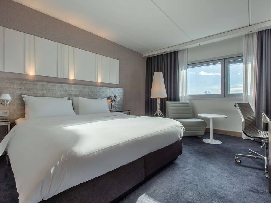 A modern hotel room with a large bed, a desk, a chair, and a window with curtains.