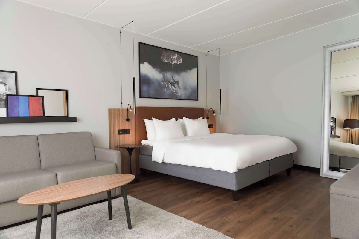 A modern hotel room with a bed, a sofa, a small table, and a painting on the wall.