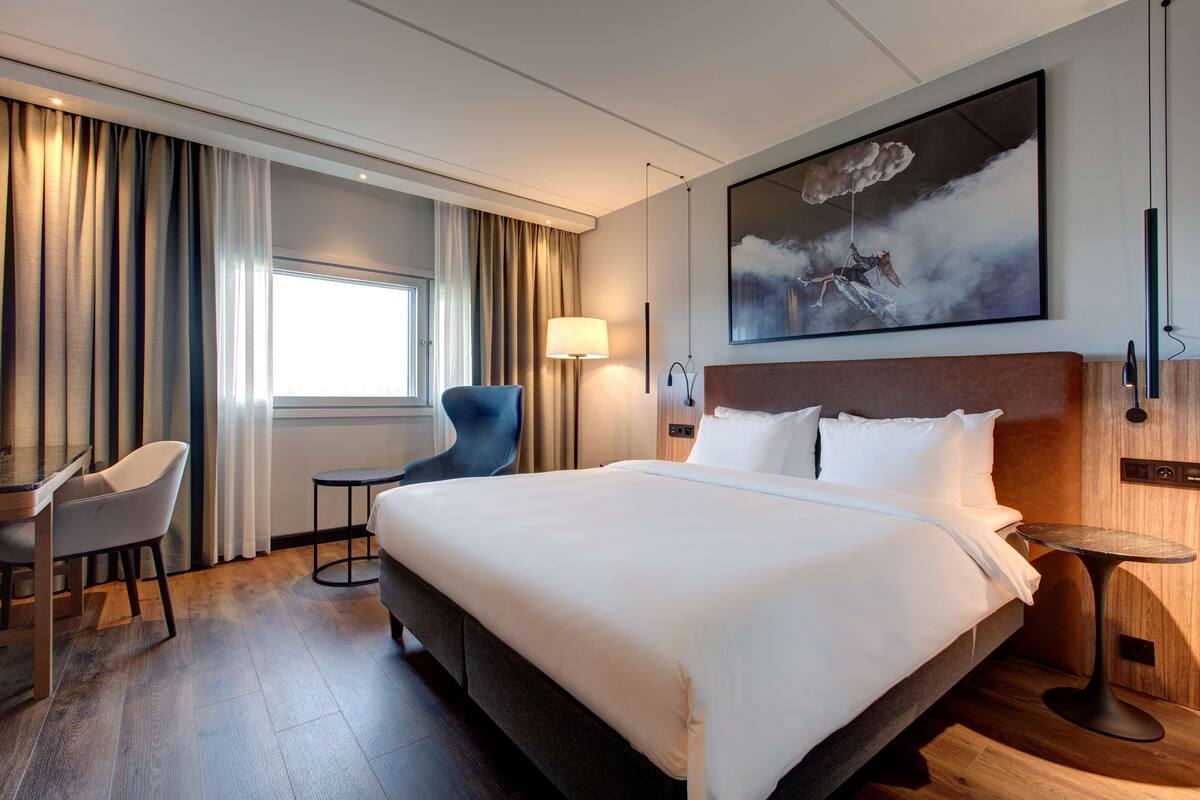 A hotel room with a large bed, a desk, a chair, a small table, and a painting on the wall.