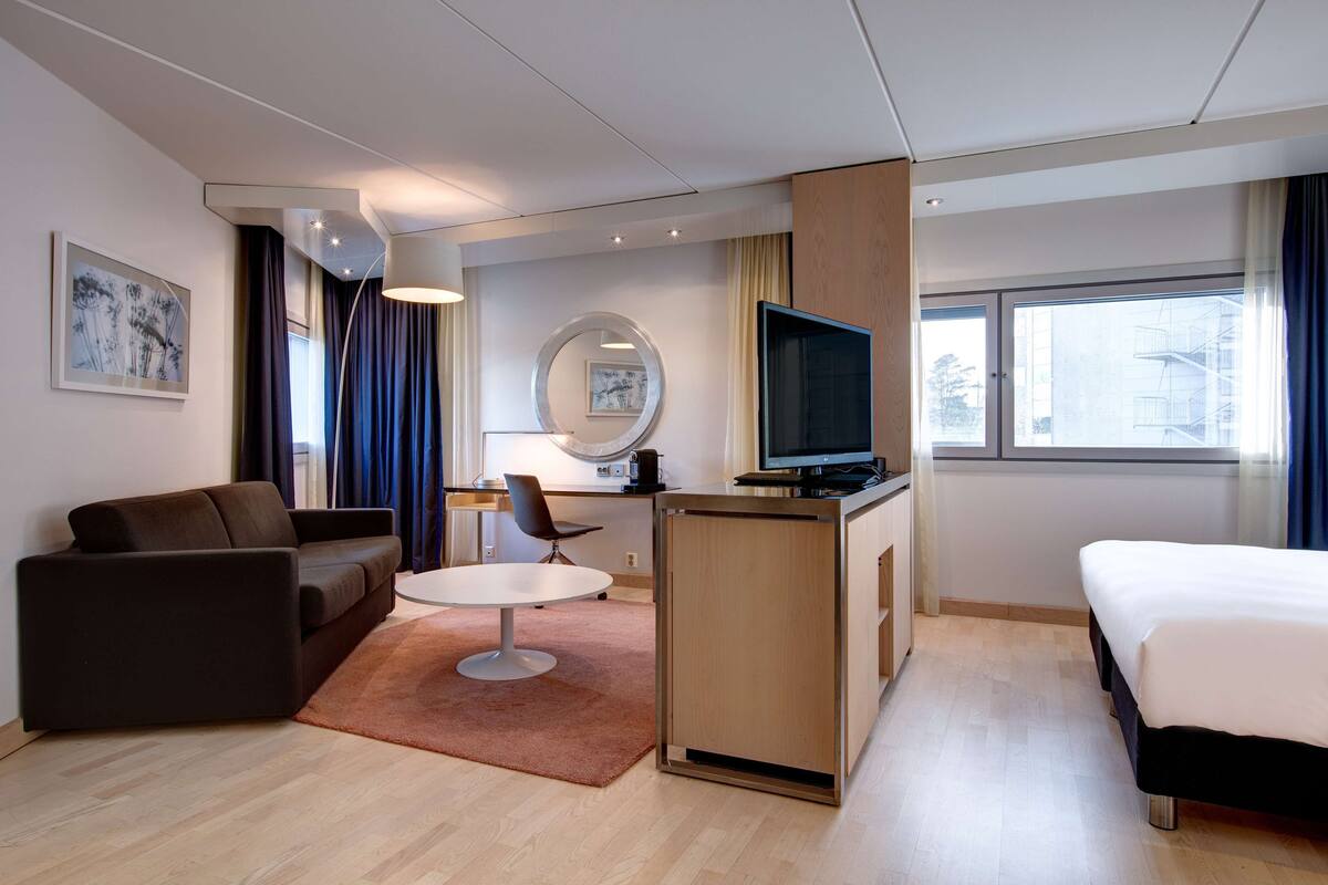 A modern hotel room with a sofa, a desk with a chair, a bed, and a TV.