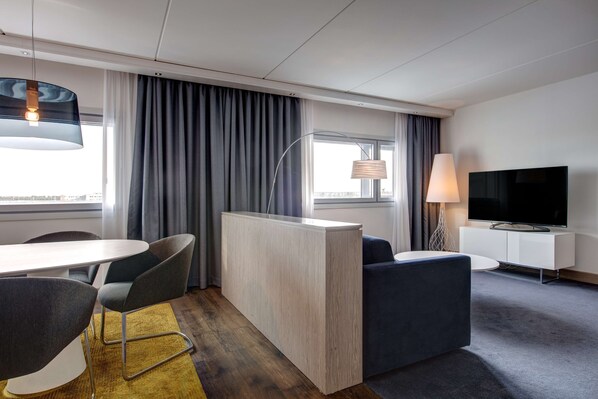 A modern hotel room with a dining area, a sofa, a TV, and a large window with curtains.