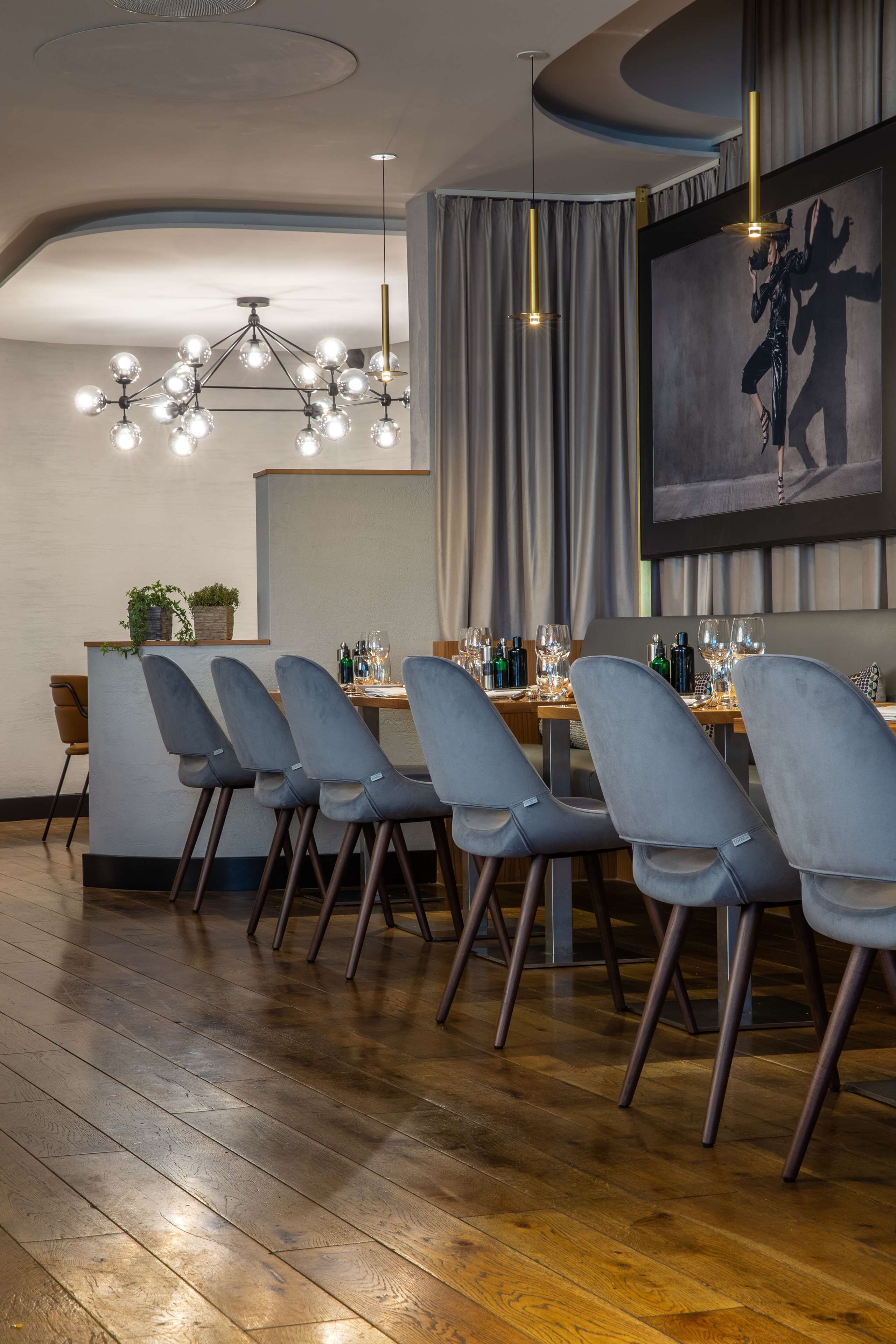 A dining area with a long wooden table, grey upholstered chairs, and a modern chandelier.