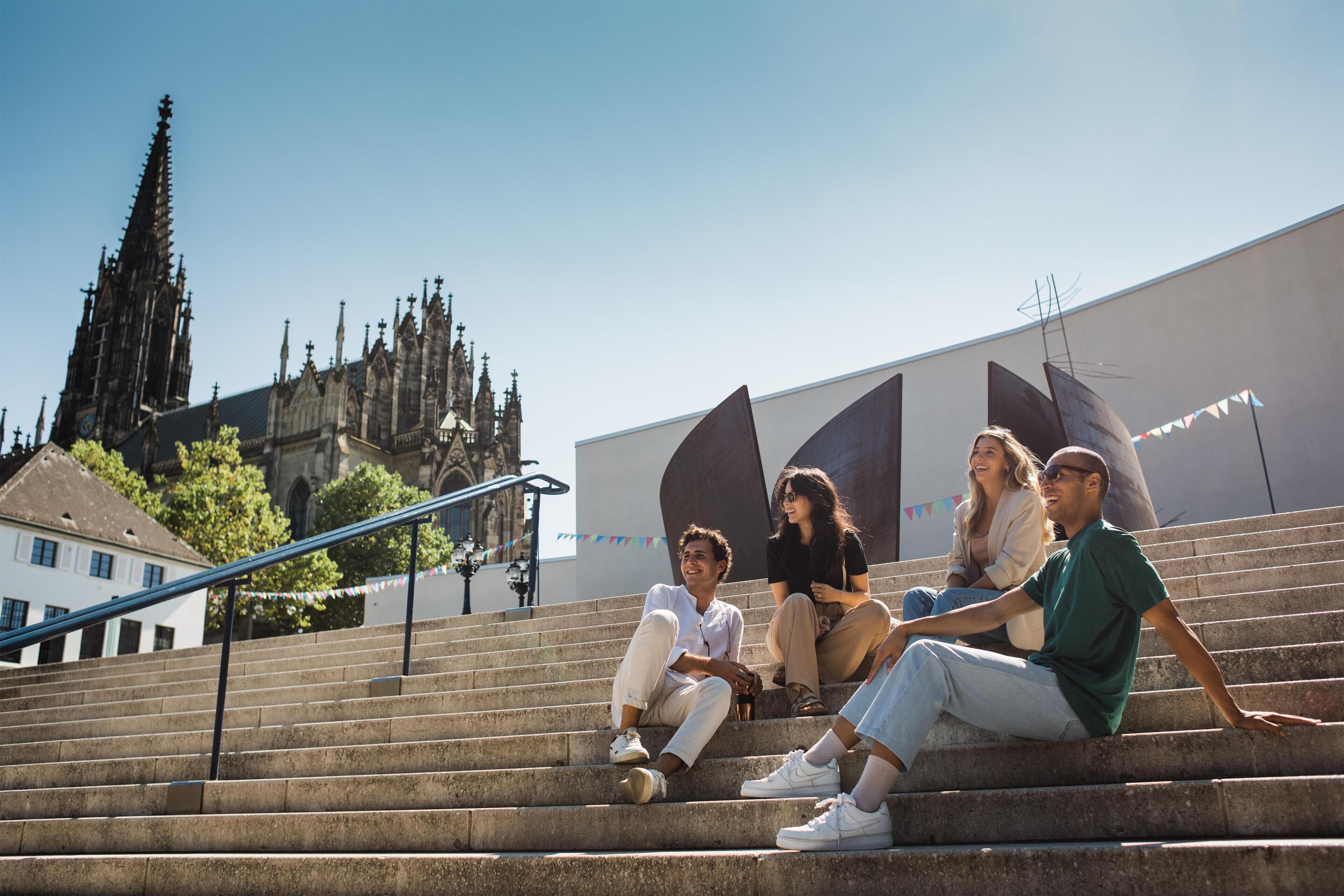 A group of people sitting on outdoor stairs with a historic building in the background.