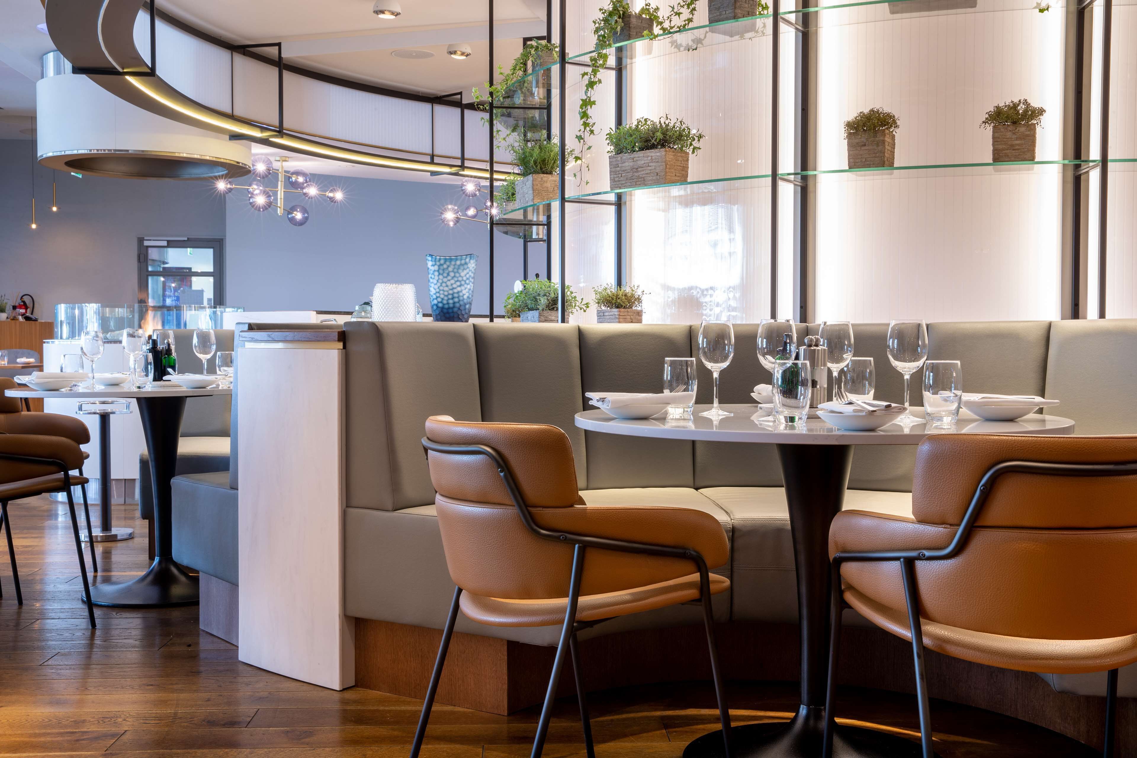 Un restaurante moderno con una zona de estar curvada, estantes de vidrio con plantas y una barra.