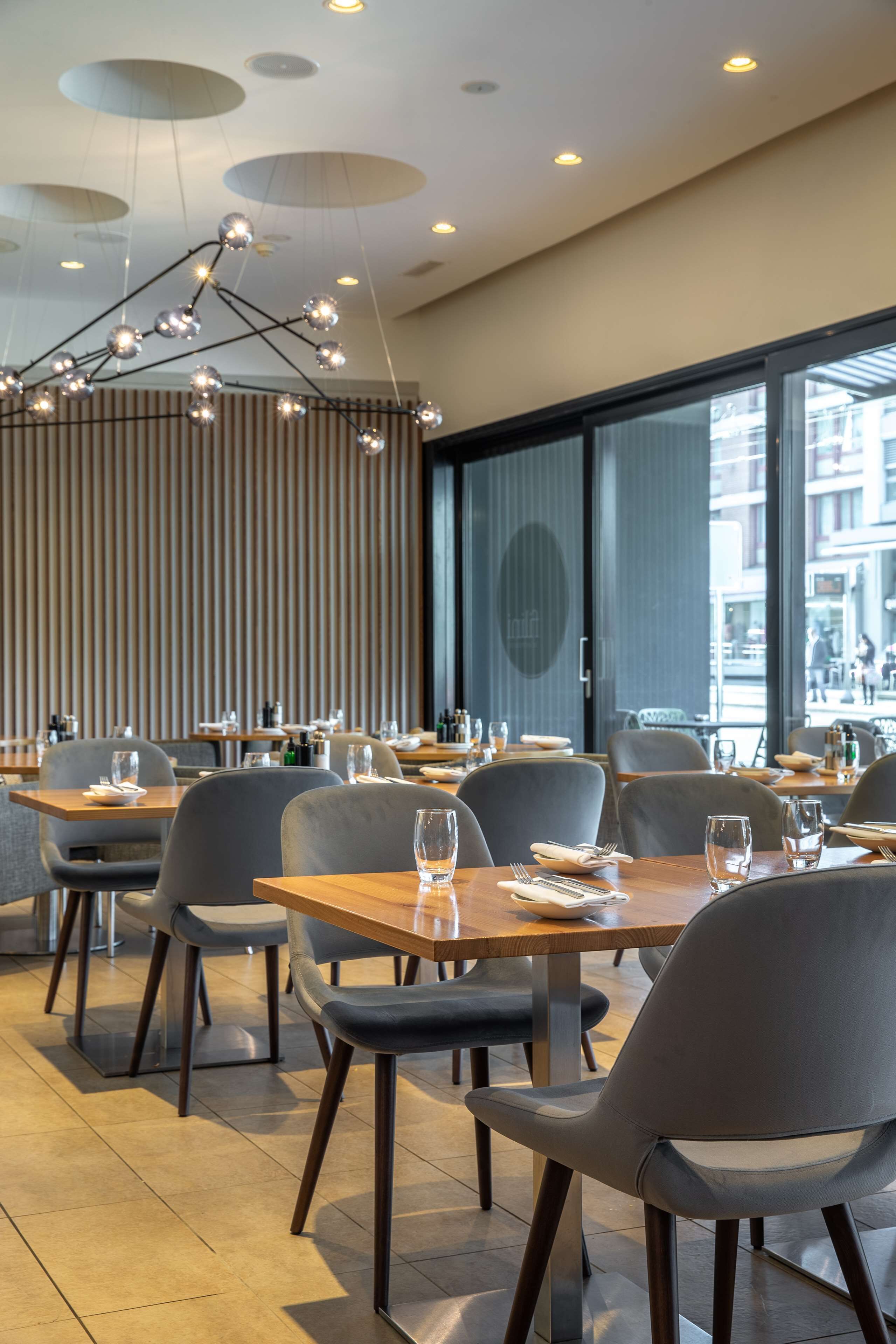 A modern restaurant interior with wooden tables and grey chairs, a unique ceiling light fixture, and large windows offering a view of the street.