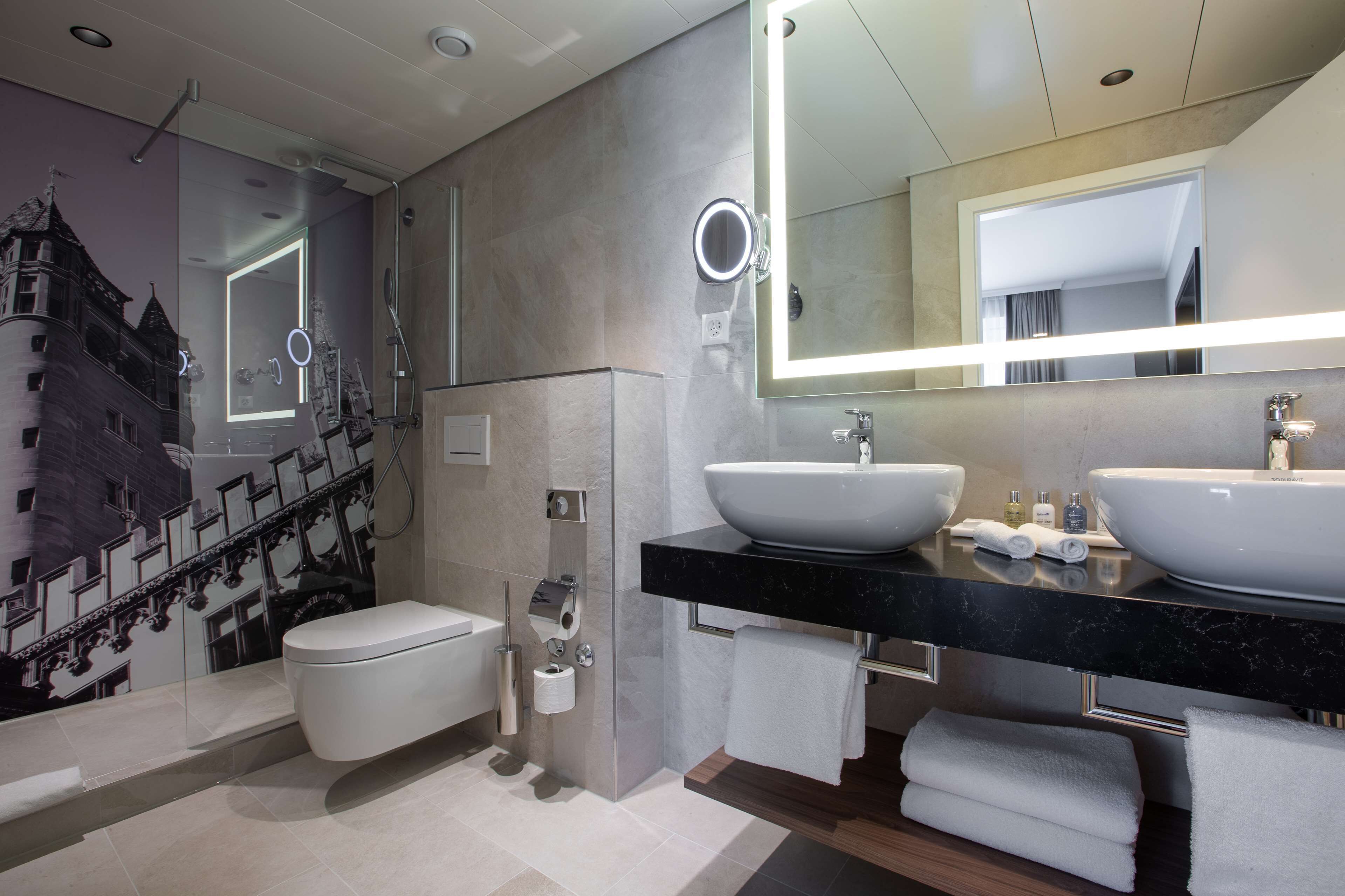 A modern bathroom with a large mirror, two sinks, and a toilet.