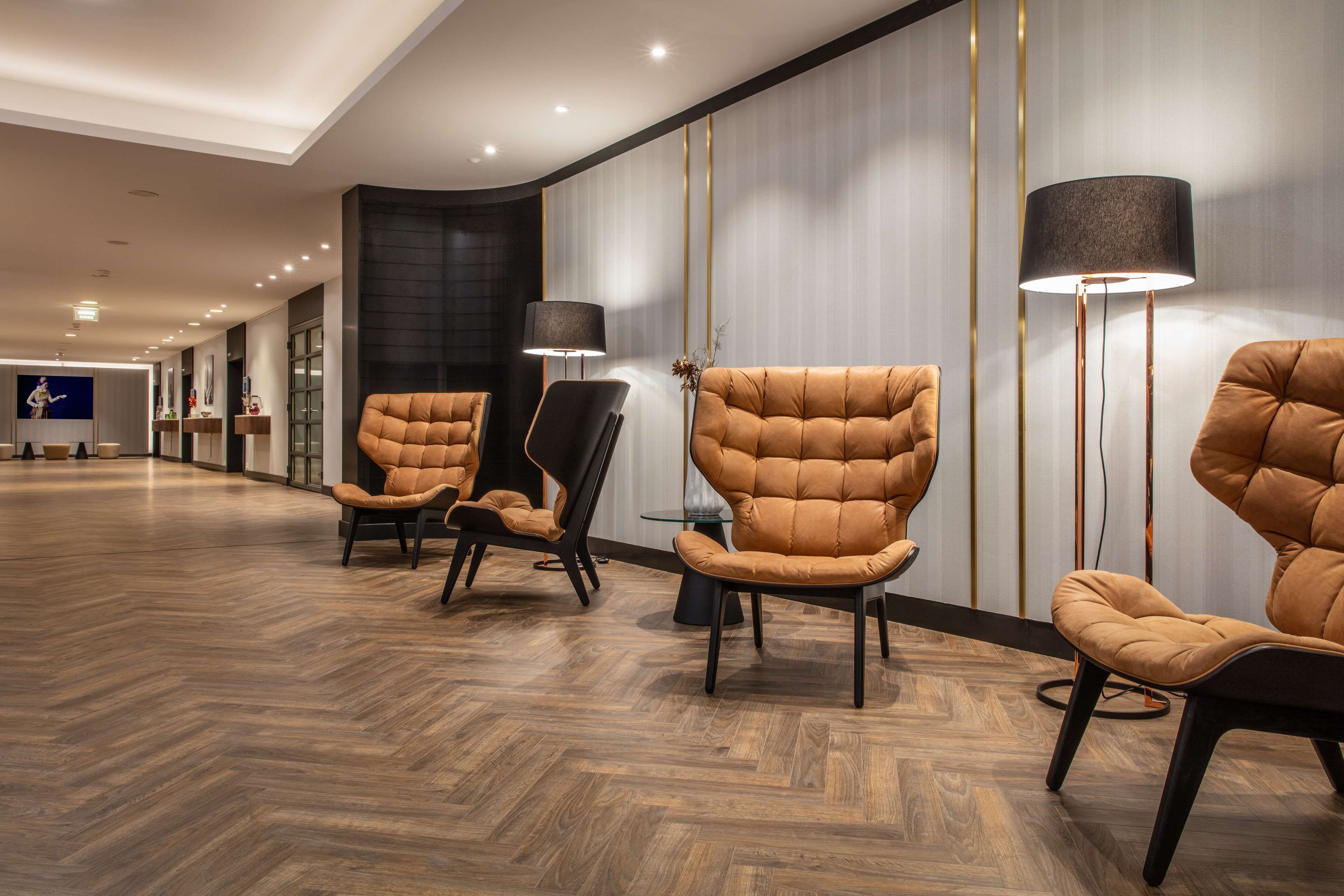 A modern hotel lobby with a wooden herringbone floor, leather armchairs, a standing lamp, and a wall-mounted TV.