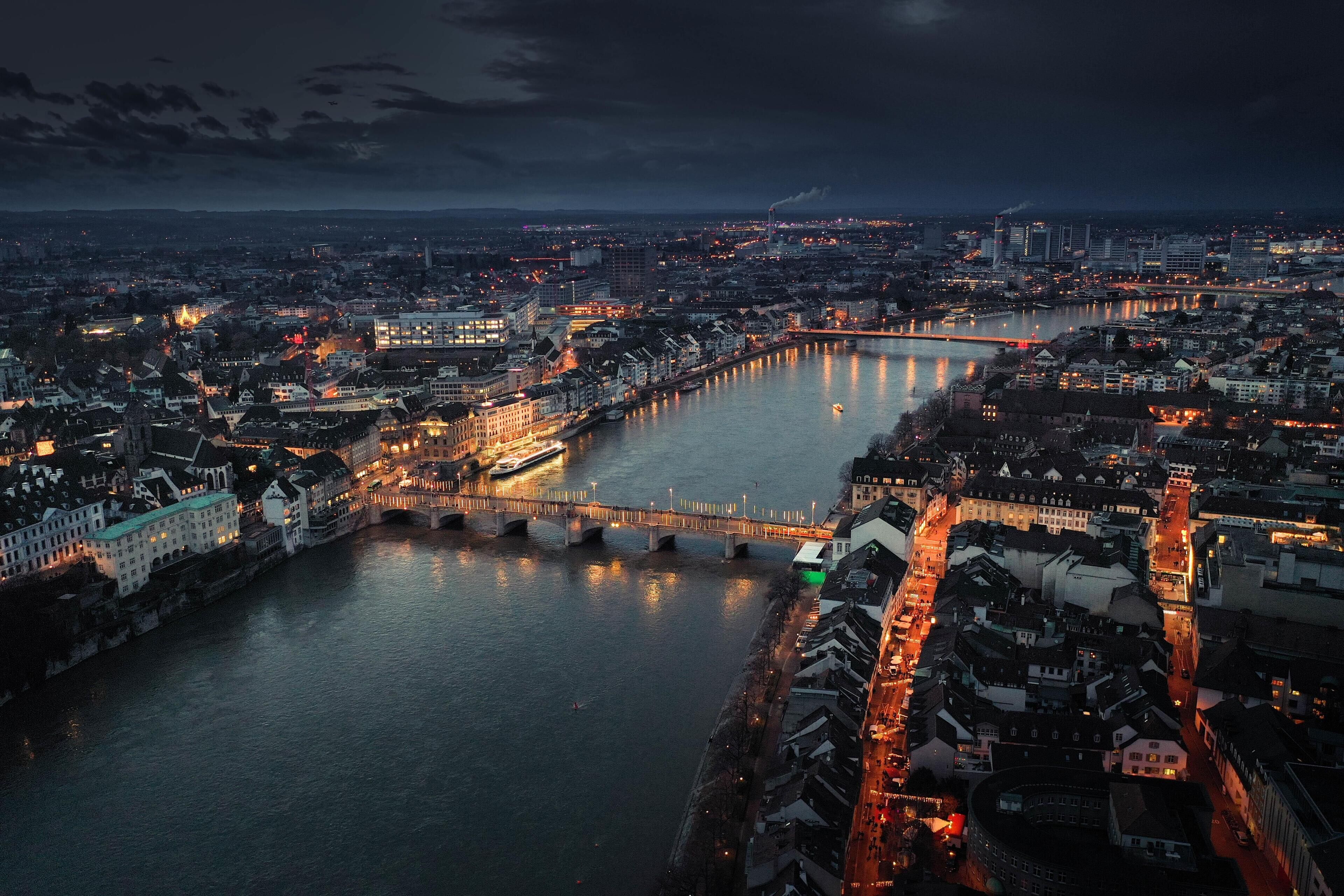 An aerial view of a cityscape at dusk with a river, bridges, and illuminated buildings.
