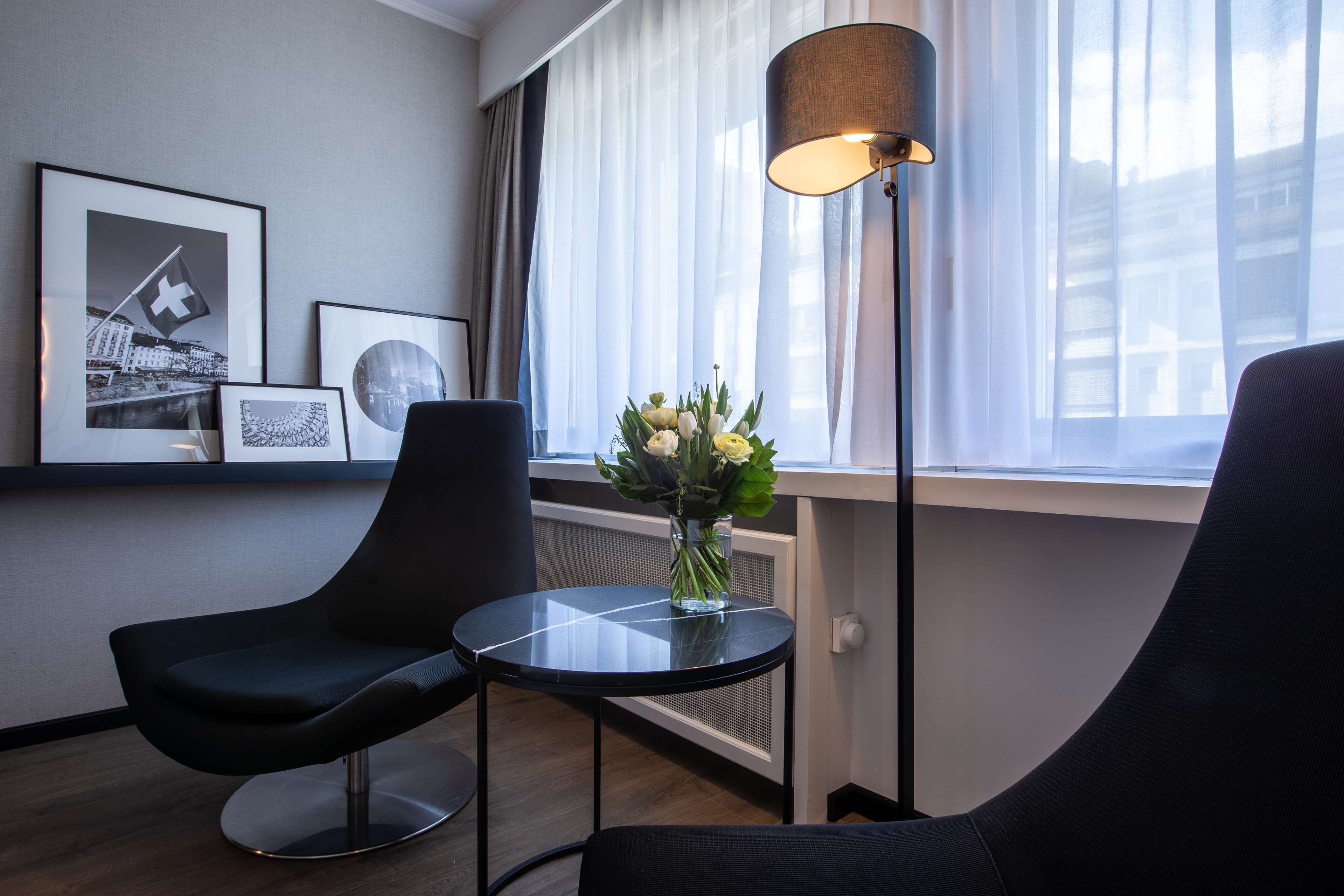 Een moderne hotelkamer met twee zwarte stoelen, een klein rond tafeltje, een vloerlamp en een vaas met bloemen. Er hangen afbeeldingen in lijsten aan de muur.