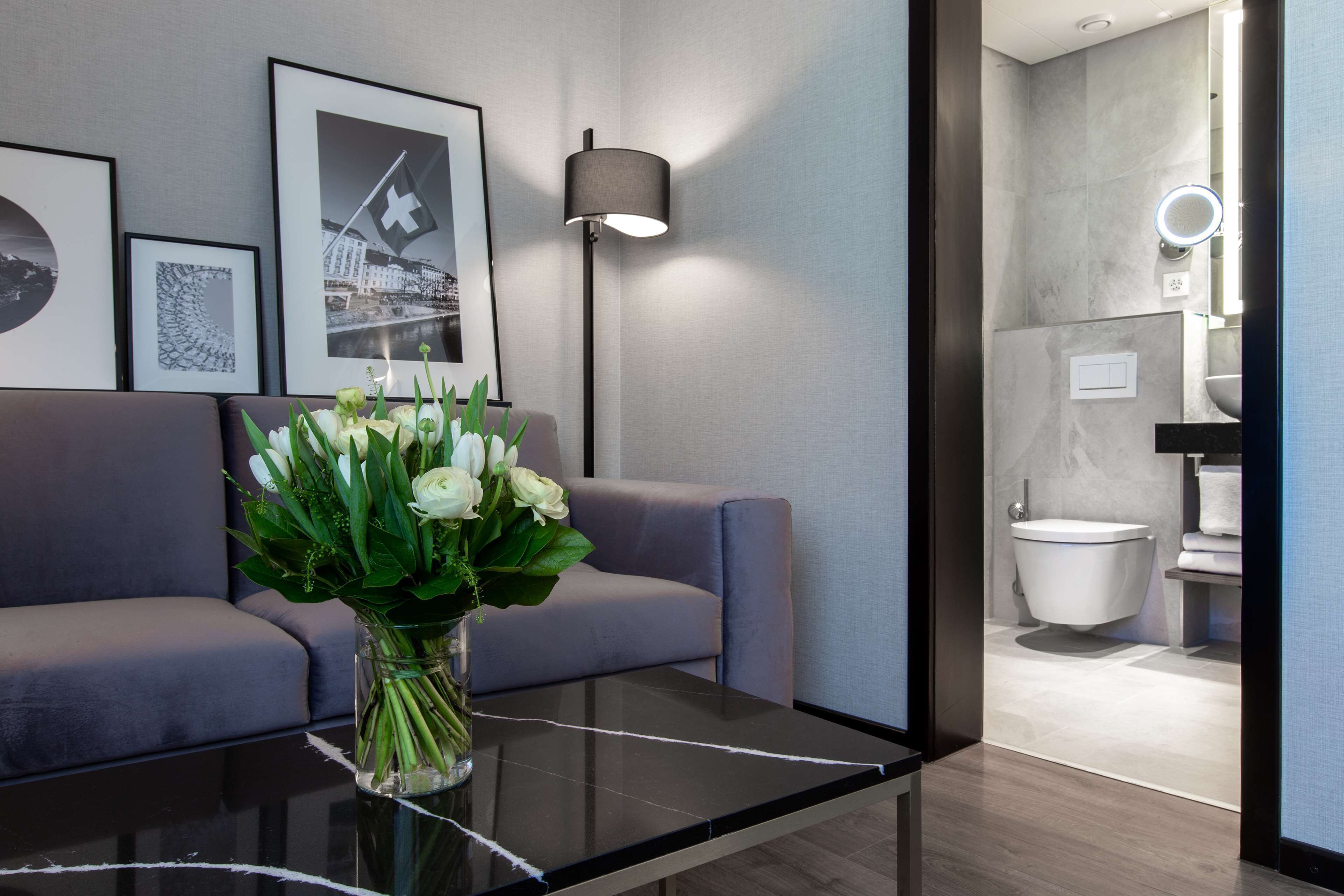 Un salon moderne avec un canapé gris, une table basse en verre ornée d’un vase de fleurs blanches et des tableaux encadrés accrochés au mur.
