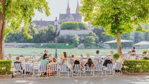Un área de comedor junto al río con mesas al aire libre, vista a un edificio histórico y un puente al fondo.
