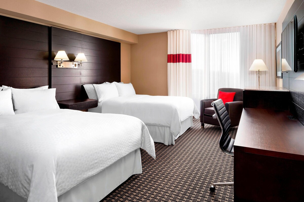 A hotel room with two beds, a desk, a chair, a TV, and a window with curtains.