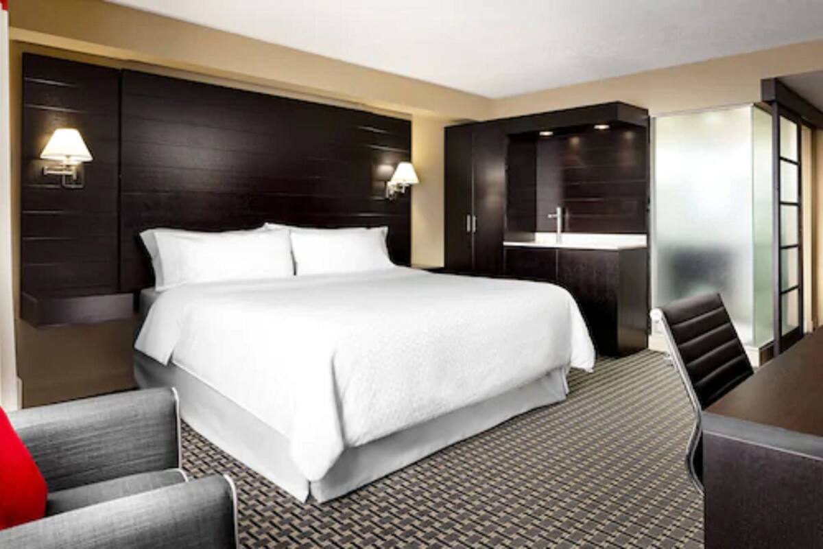 A hotel room with a large bed, a desk, and a chair. There are two wall-mounted lamps and a minibar.