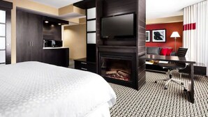 Habitación de hotel moderna con una cama grande, un televisor de pantalla plana montado en una pared oscura, una chimenea, un escritorio con una silla y una pared de acento roja.