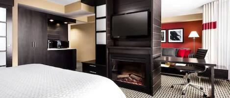 A modern hotel room with a large bed, a flat-screen TV mounted on a dark wall, a fireplace, a desk with a chair, and a red accent wall.