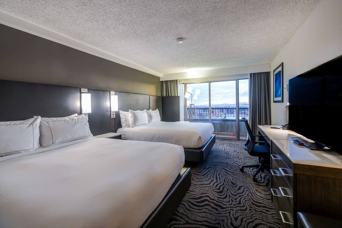 A hotel room with two beds, a desk, a chair, a TV, and a large window with a city view.