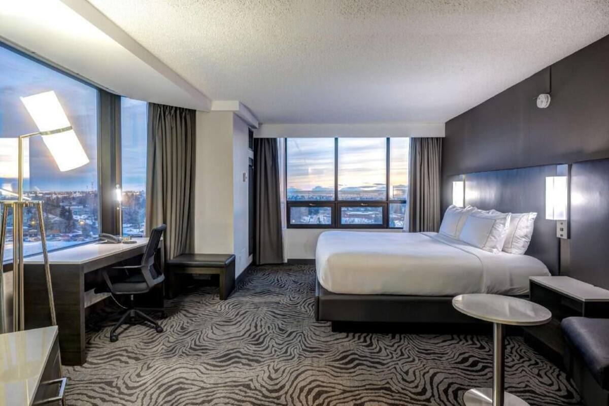 A hotel room with a large bed, a desk, a chair, a small table, and a view of the city through large windows.