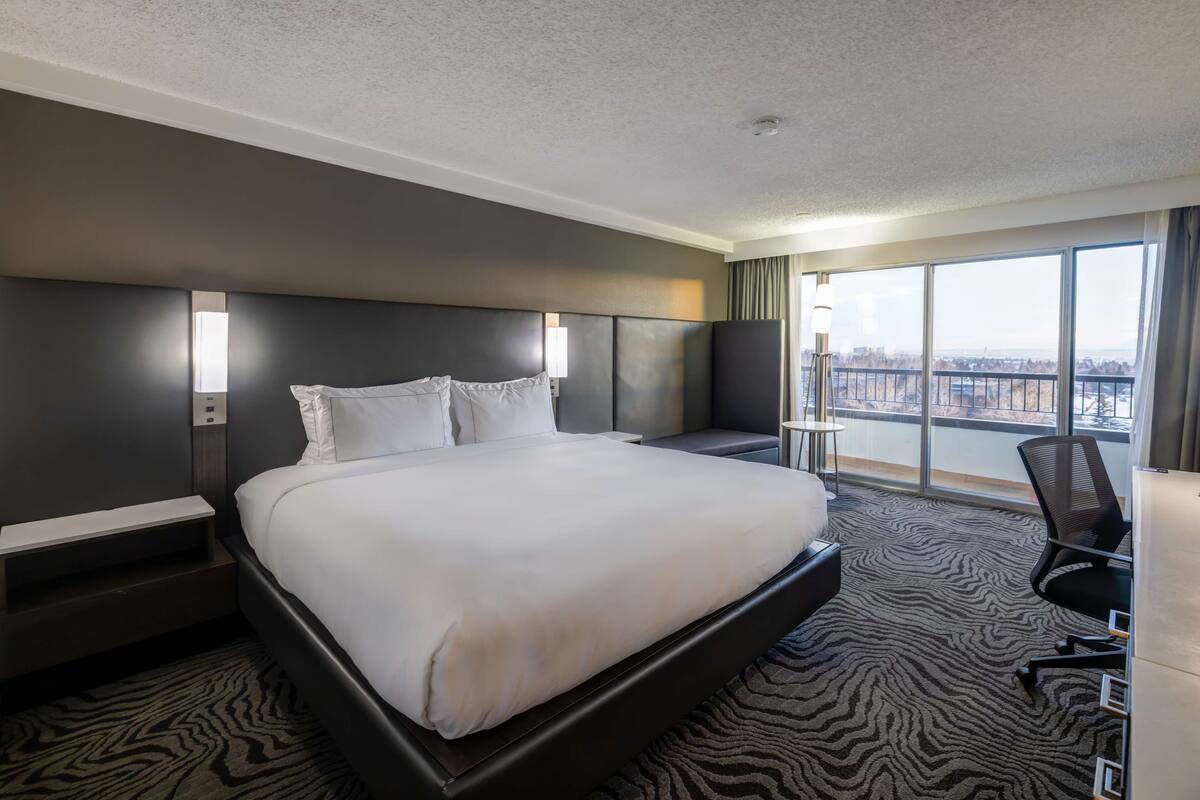 A hotel room with a large bed, a desk, a chair, and a view of the city through large windows.