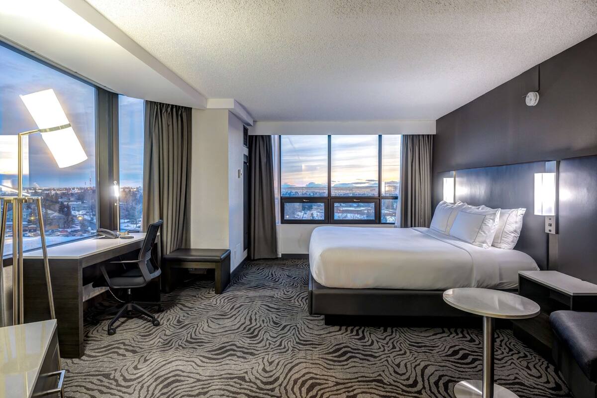A hotel room with a large bed, a desk, a chair, a small round table, and a view of the city through large windows.