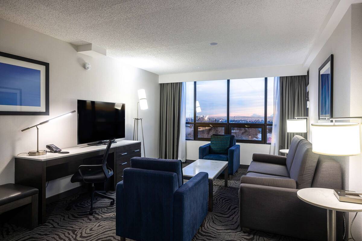 A hotel room with a desk, a television, a sofa, and a view of the city.