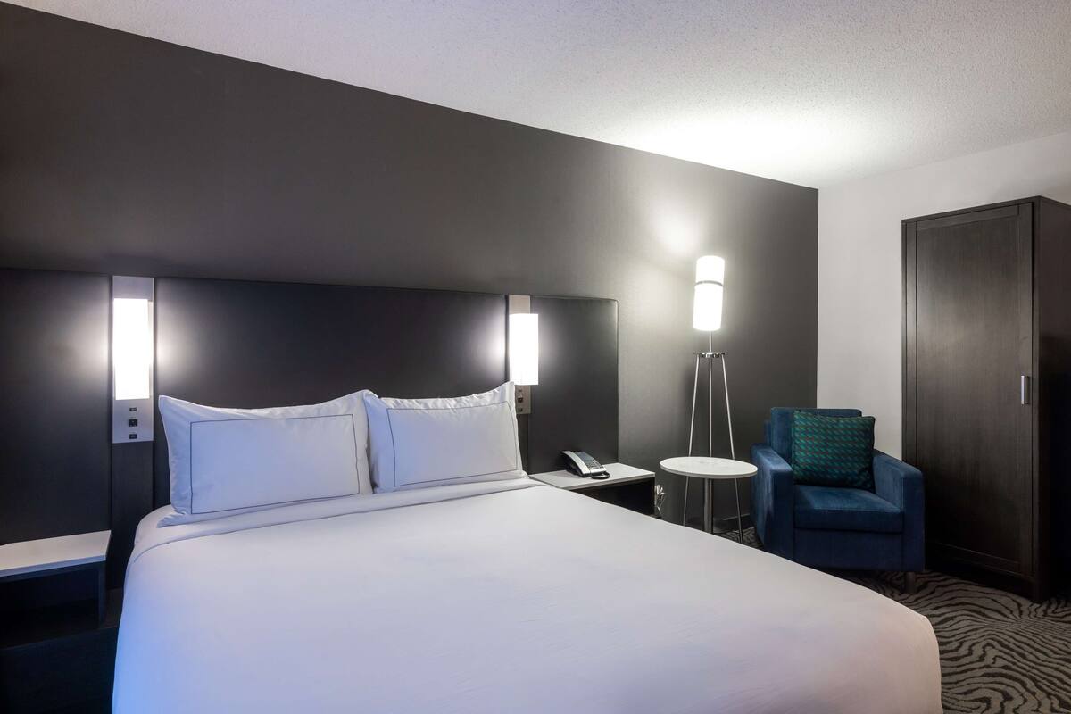 A hotel room with a large bed, bedside tables, a chair, a wardrobe, and a lamp.