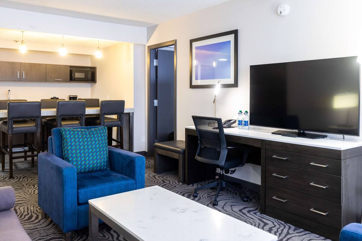 A modern hotel room with a dining area, a large flat-screen TV, a comfortable sofa, and a desk with drawers.