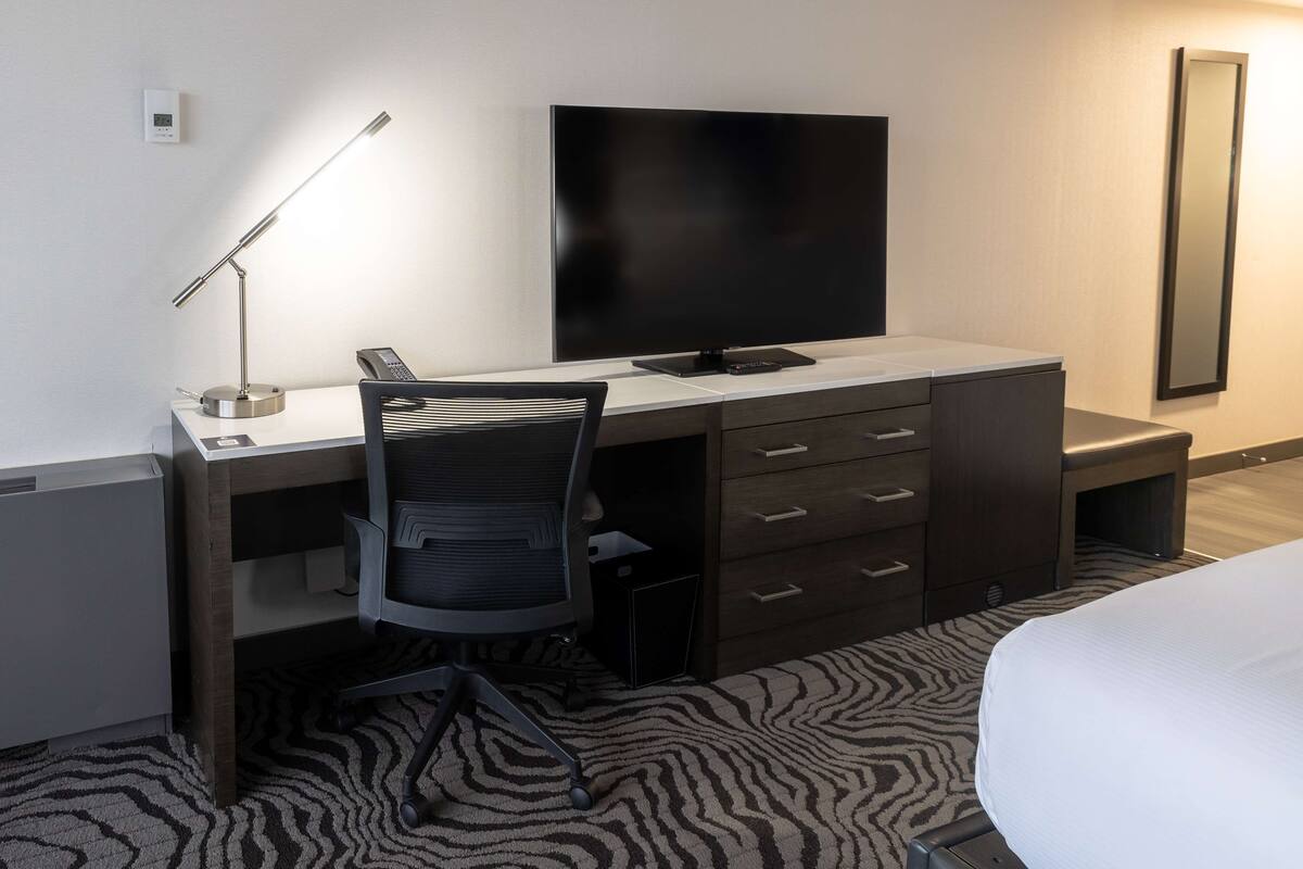 A hotel room with a bed, a desk with a computer, a chair, a flat-screen TV, a mirror, and a patterned carpet.