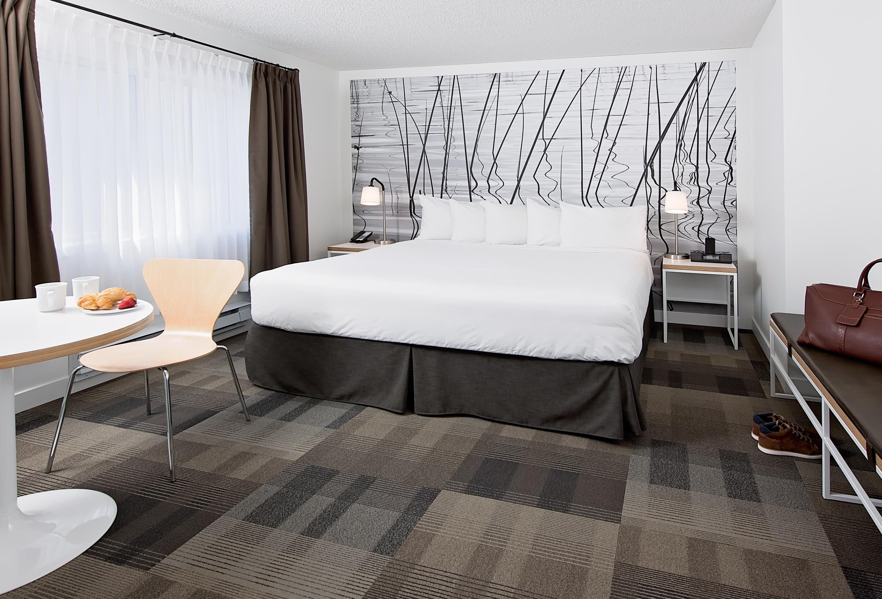 A hotel room with a large bed, a desk with a chair, a small table with pastries, and a handbag on a bench.