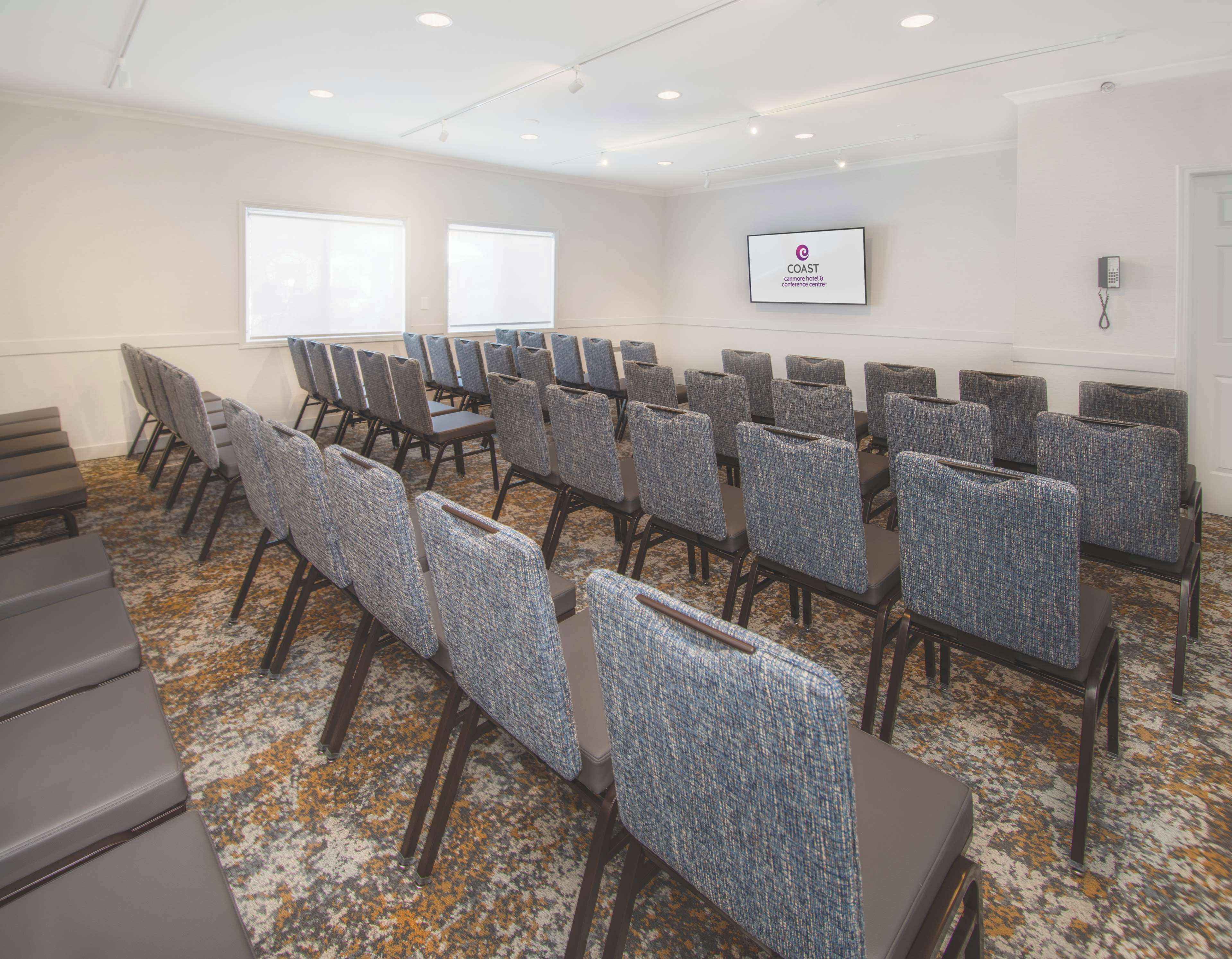 Una sala de conferencias con filas de sillas, piso alfombrado, una pantalla en la pared y un teléfono.