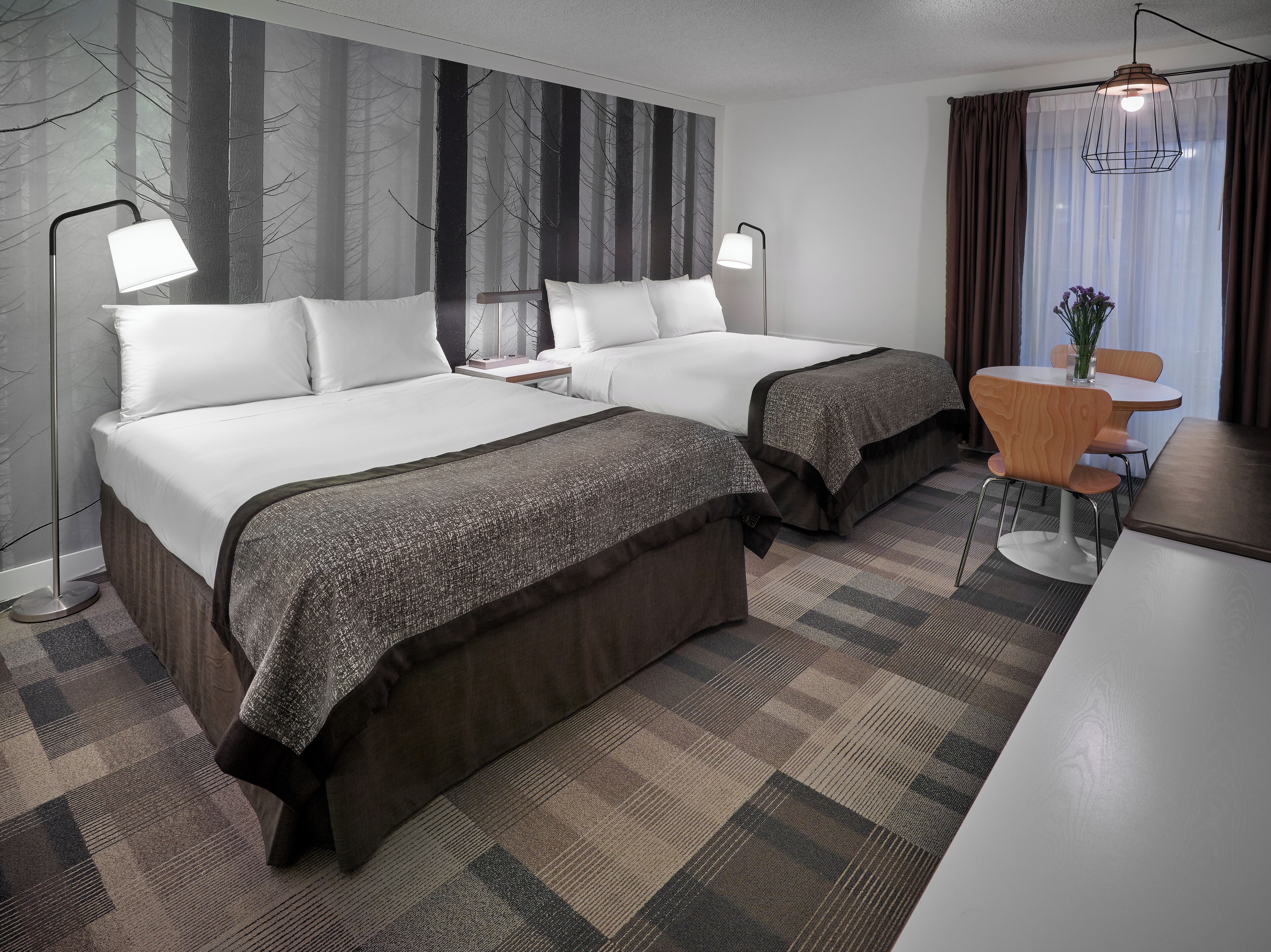 Quarto de hotel com duas camas, uma escrivaninha, uma cadeira e uma janela grande com cortinas.