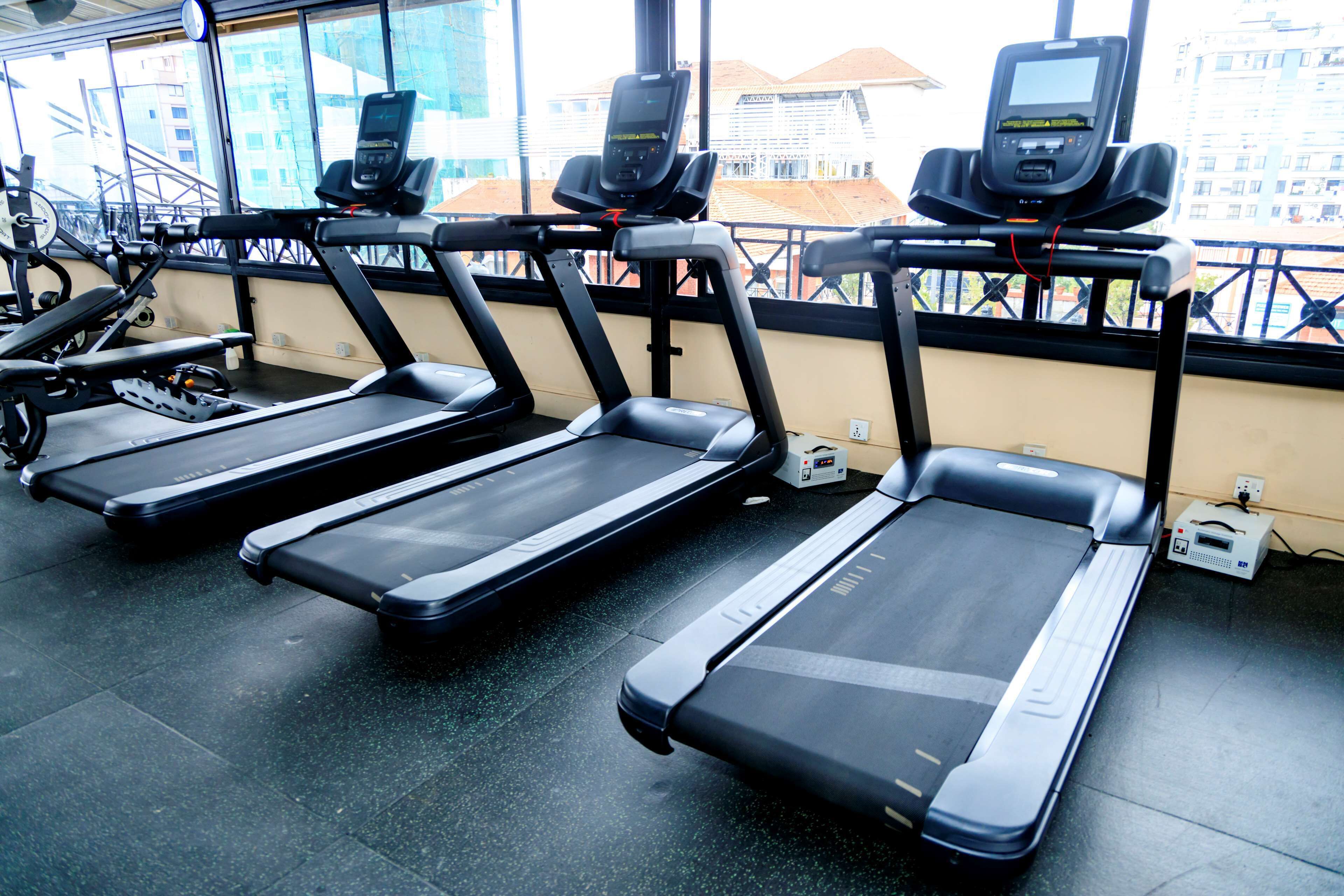 A row of stationary treadmills in a gym with large windows and a view of buildings outside.
