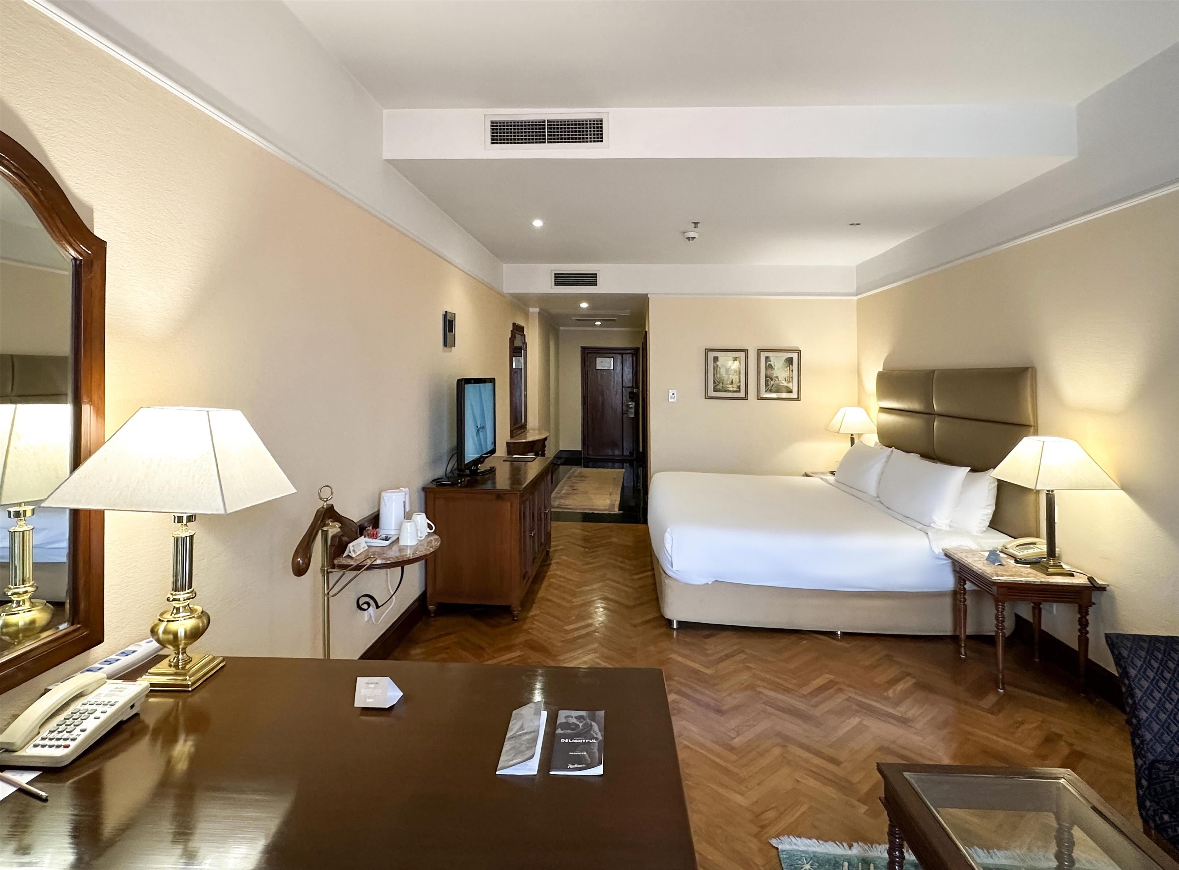 Une chambre d’hôtel comprenant un lit, des tables de chevet avec des lampes, un bureau avec un téléphone, un miroir et un téléviseur.