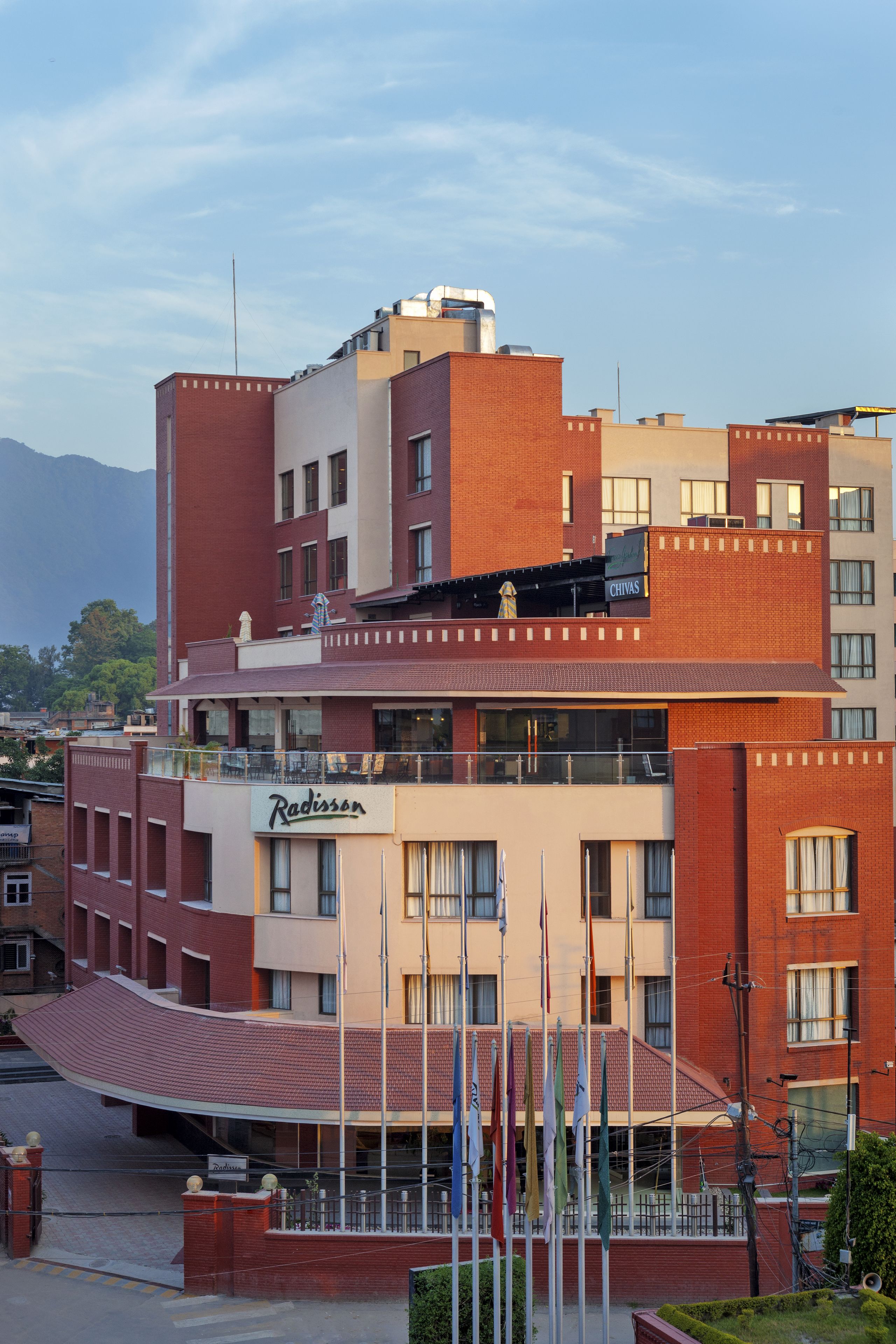 Un hotel de varios pisos con un llamativo letrero de "Radisson", que presenta un diseño arquitectónico moderno con ladrillo rojo y detalles color crema, y un patio con banderas.