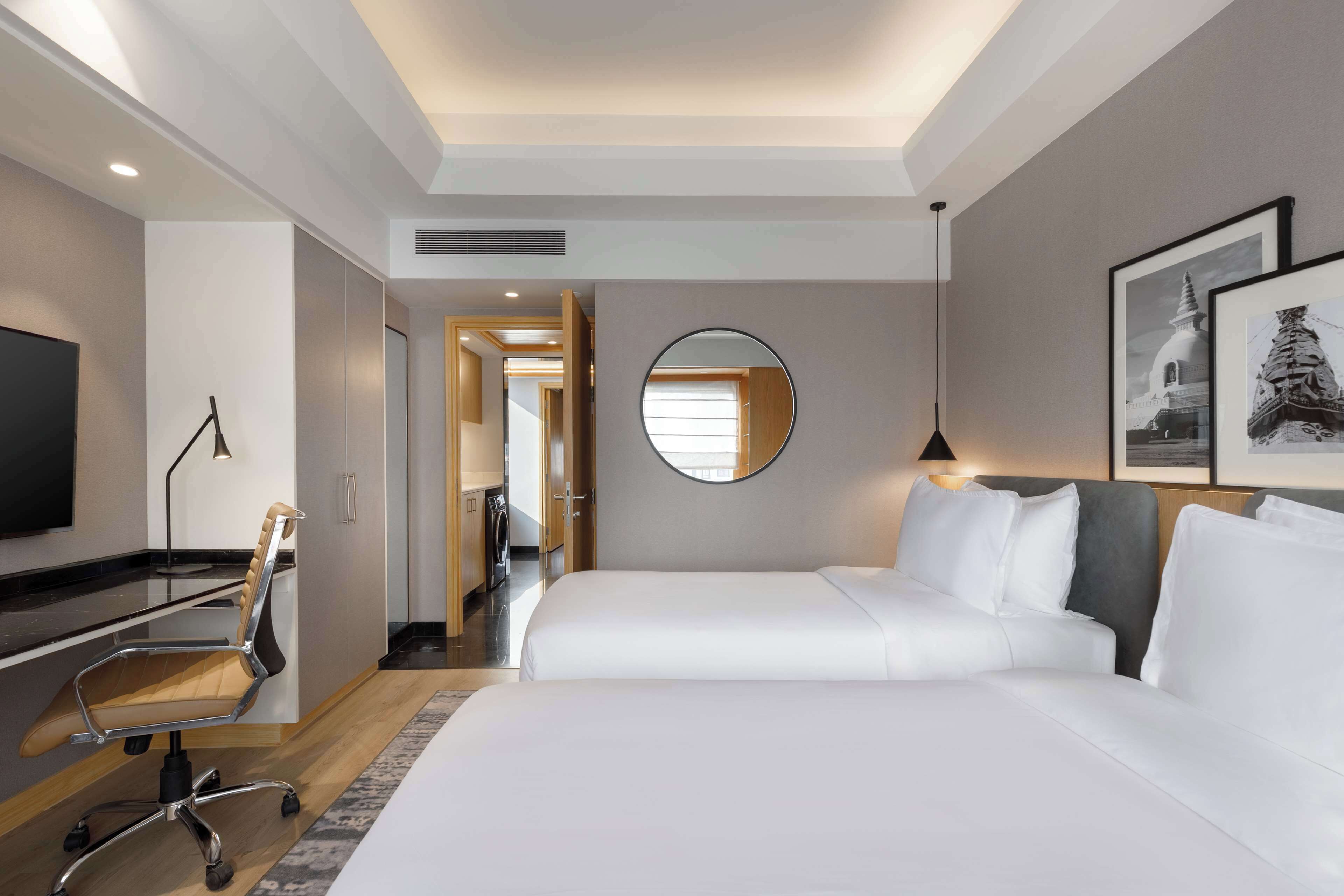 A hotel room with two beds, a desk, a chair, a TV, a mirror, and framed pictures on the wall.