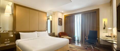 A hotel room with a large bed, a desk, a chair, a small table, and a large window with curtains.
