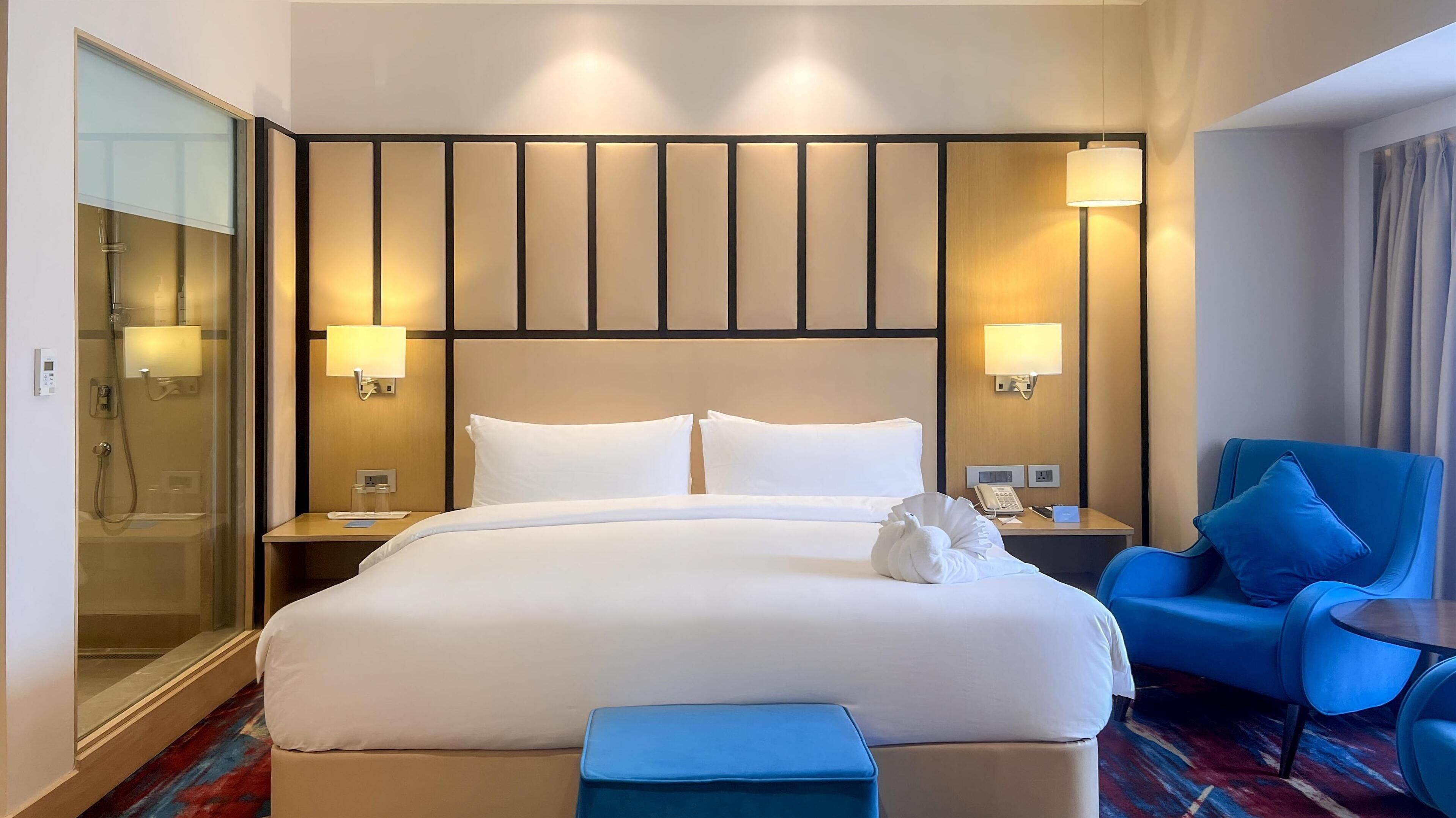 Habitación de hotel con una cama grande, dos lámparas de noche, un sillón azul, una mesita y una zona de ducha con paredes de vidrio.