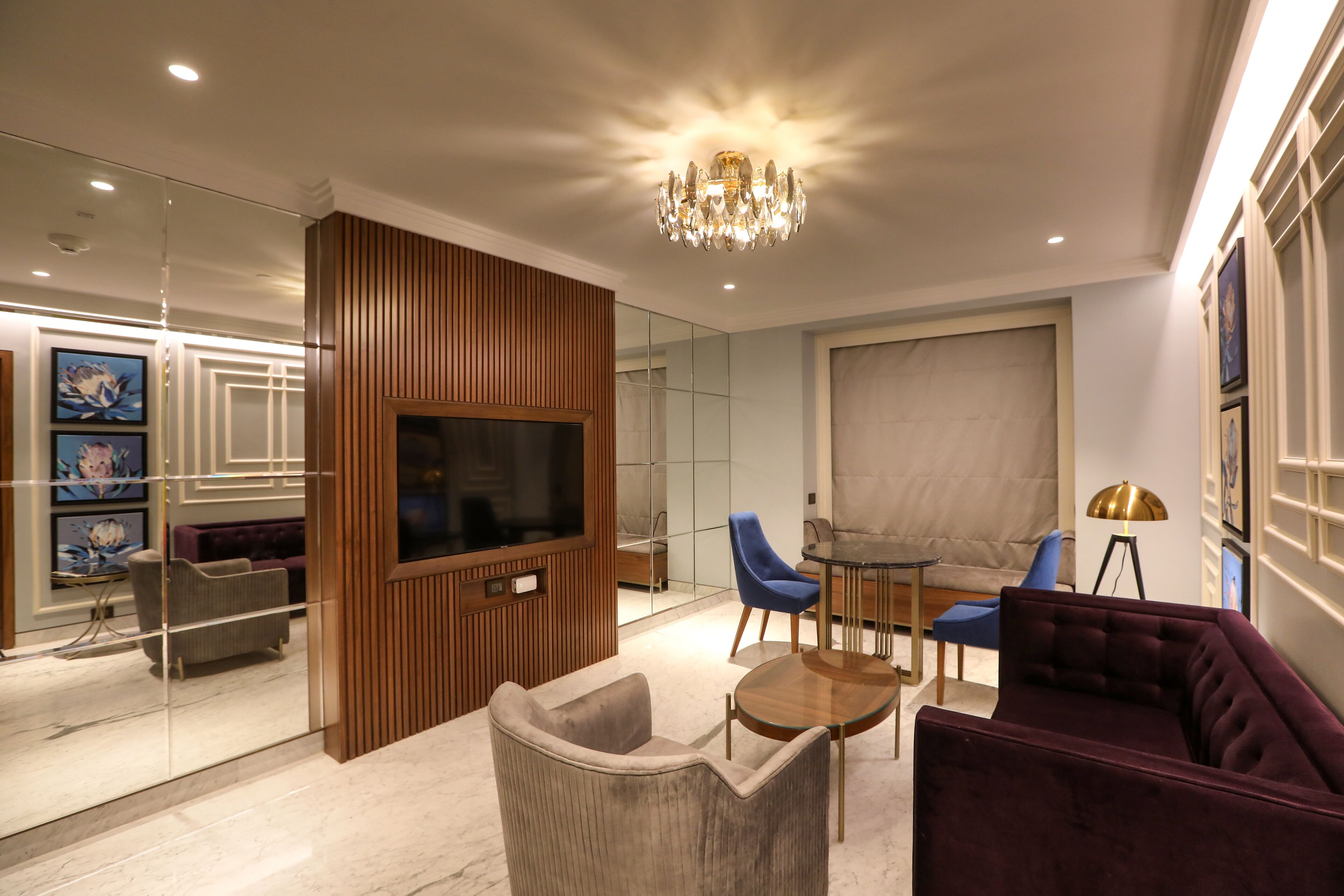A modern living room with a flat-screen TV, wooden paneling, a chandelier, and blue chairs.