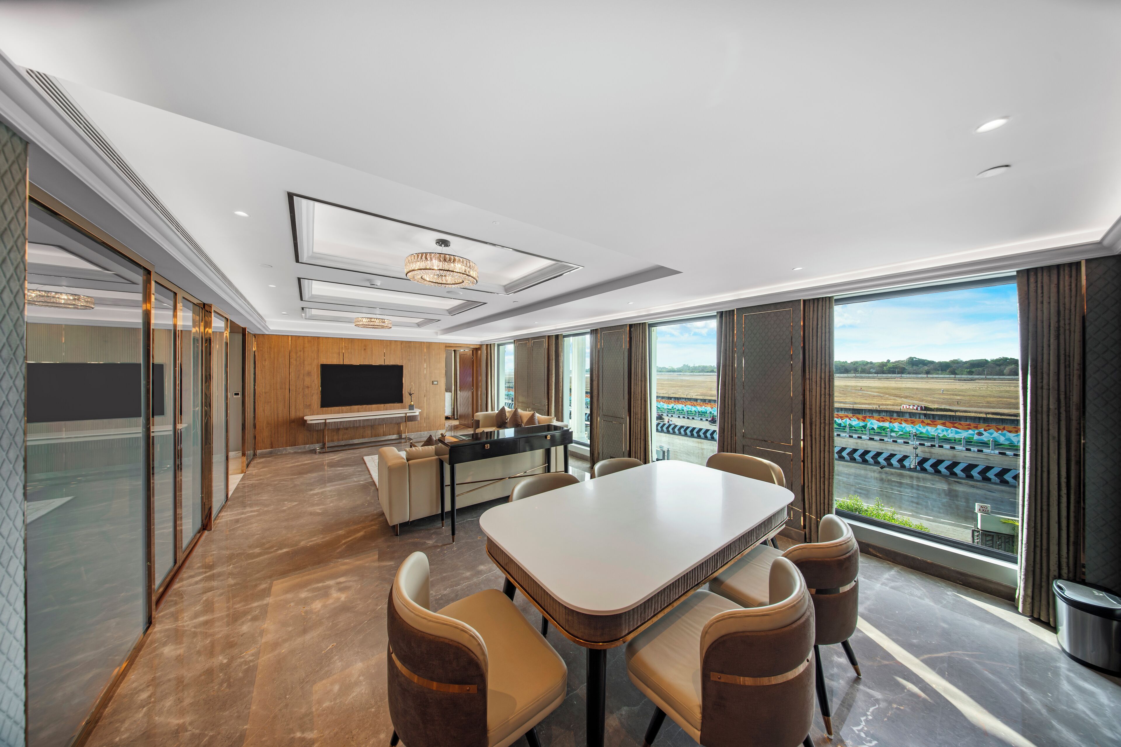 Una habitación de hotel moderna con una gran mesa de comedor, asientos cómodos, una zona de cocina y vistas a una pista de carreras.