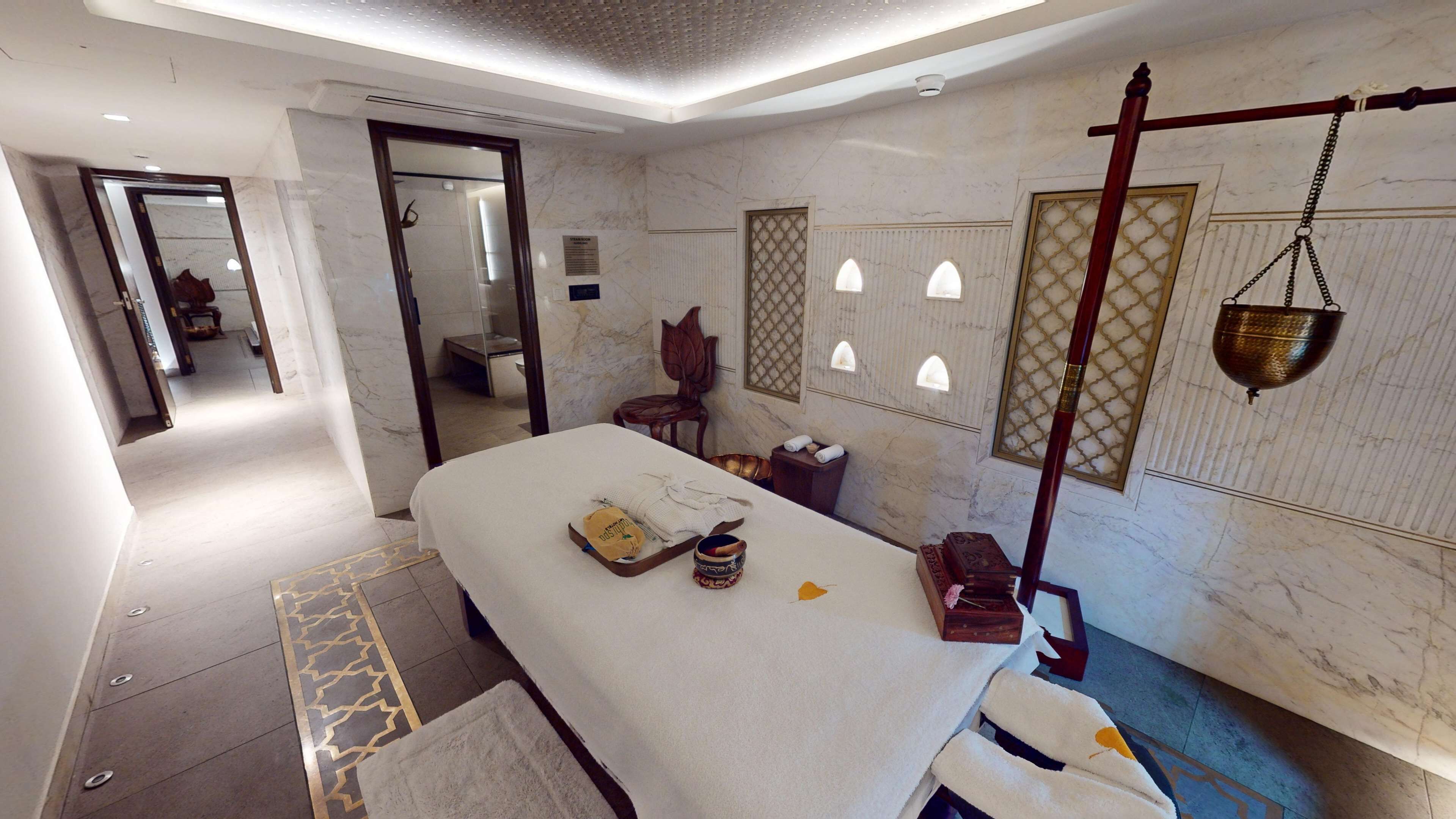 A modern spa room with a massage table, towels, a wooden chair, and a hanging bath basin.